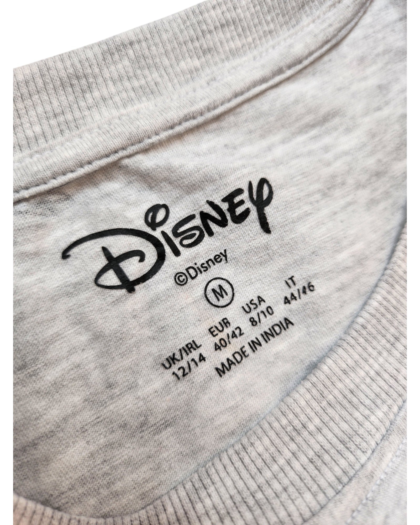 A close-up of a gray touched out mama Disney t-shirt label shows size medium (M) for UK/IRL, EUR, USA, and IT. This upcycled nursing & pumping Stitch tee is crafted in India, blending style and sustainability.