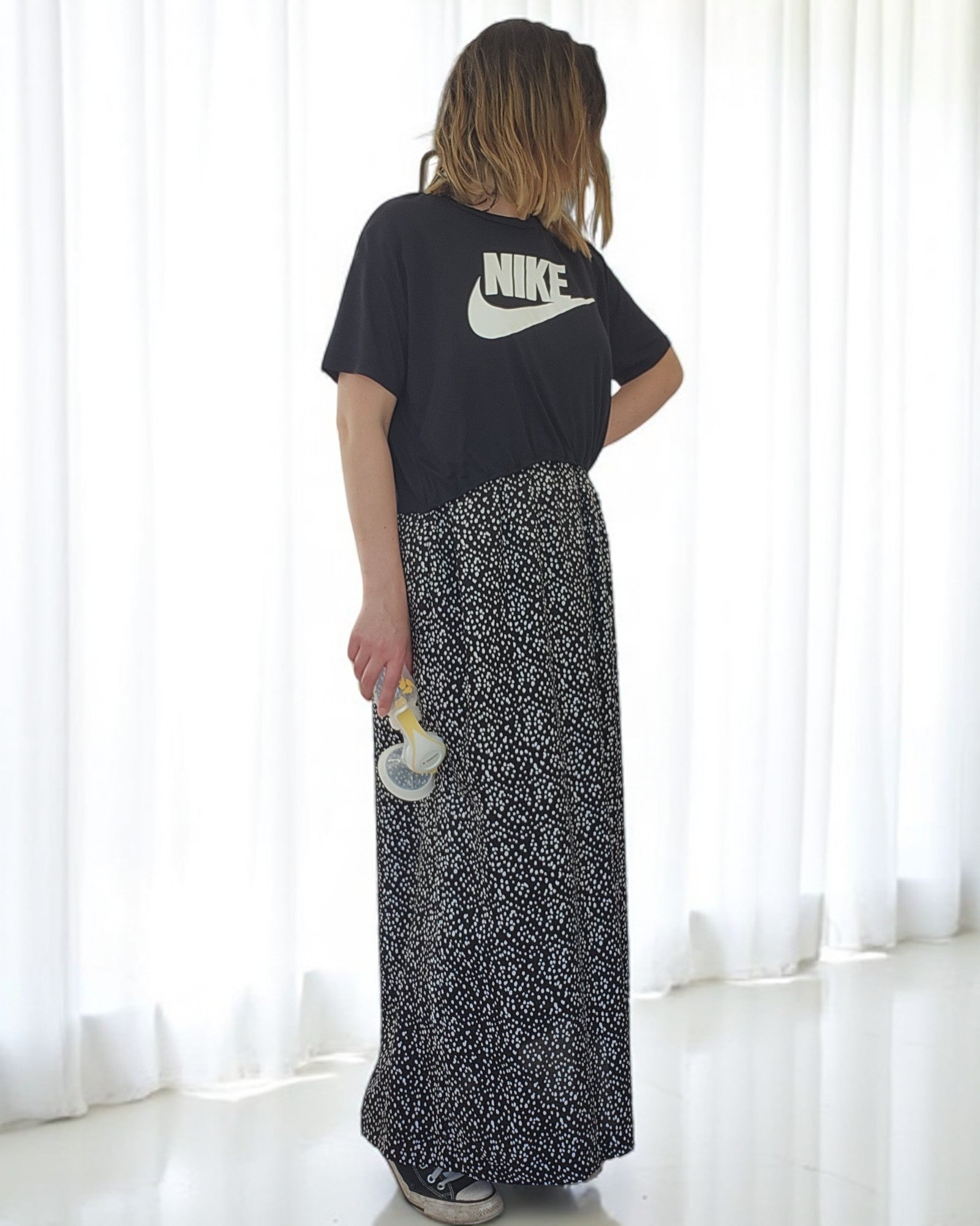 In front of white curtains, a person with shoulder-length hair wears the Upcycled Breastfeeding & Pumping NIKE Maxi Dress by touched out mama. The black and white patterned maxi dress and the breast pump emphasize the realities of nursing as their face turns away.