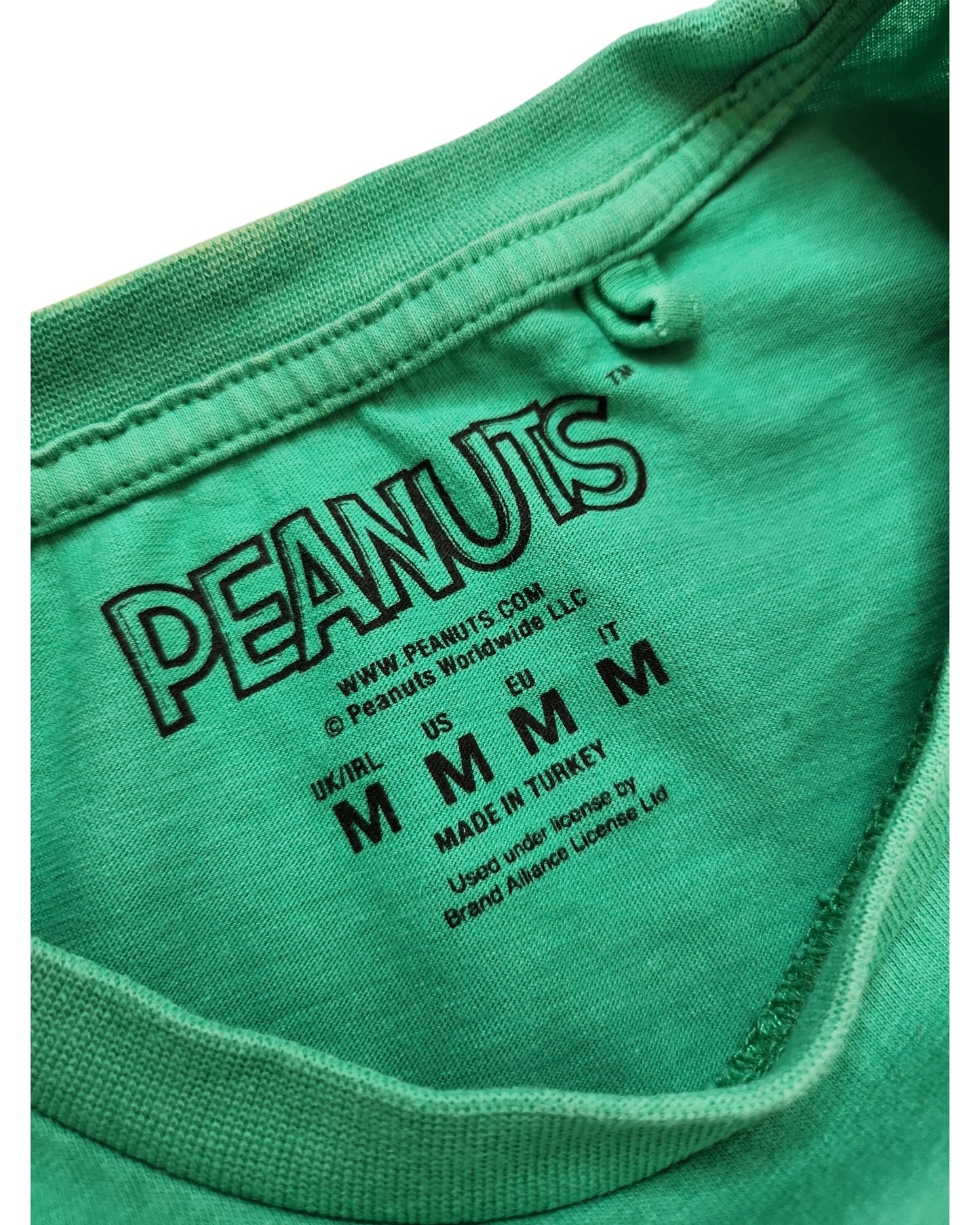 Close-up of a green Upcycled Breastfeeding & Pumping Peanuts Snoopy T-shirt label by touched out mama, crafted from eco-friendly materials. Displays logos, size info (UK/IRL/US/EU/IT: M), and made in Turkey with brand licensing details visible.