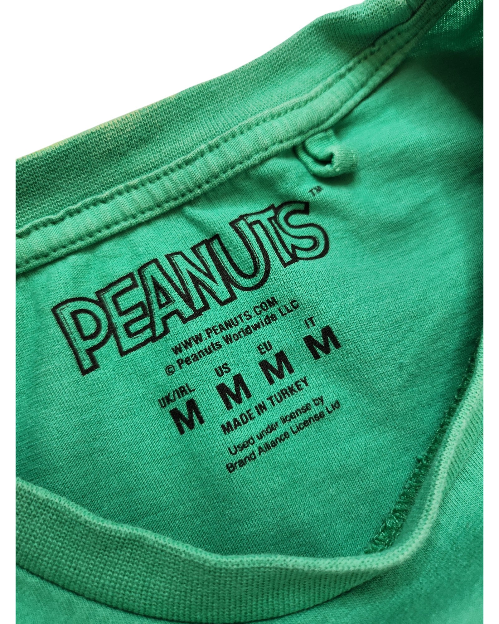 Close-up of a green Upcycled Breastfeeding & Pumping Peanuts Snoopy T-shirt label by touched out mama, crafted from eco-friendly materials. Displays logos, size info (UK/IRL/US/EU/IT: M), and made in Turkey with brand licensing details visible.