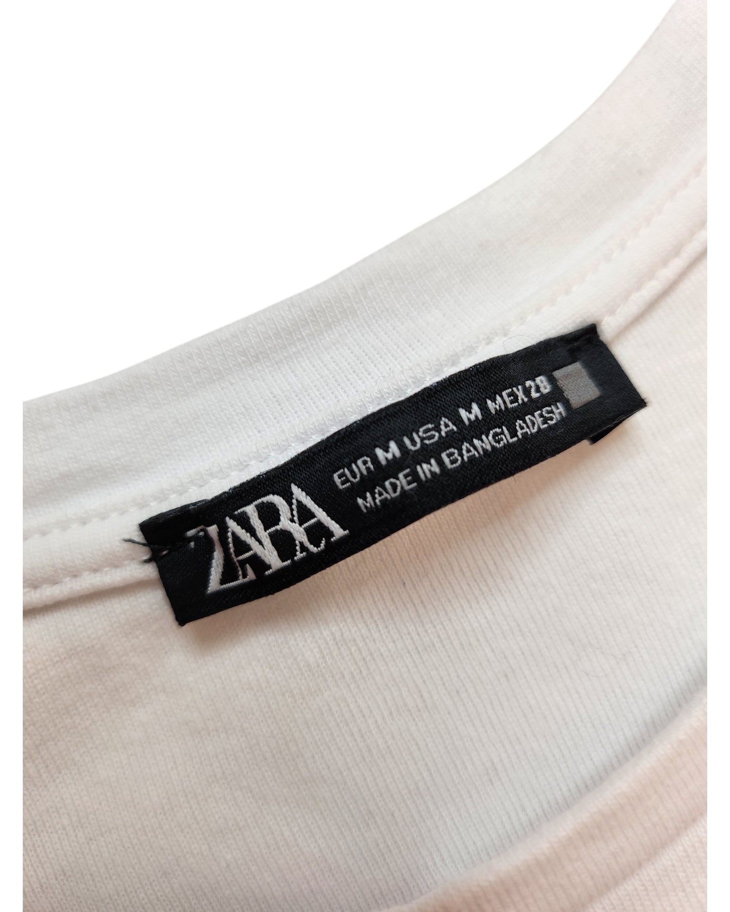 Close-up of a touched out mama label on a white ZARA t-shirt, listing sizes EUR M, USA M, MEX 28. Made in Bangladesh, this upcycled garment emphasizes eco-friendly fashion through sustainable practices.