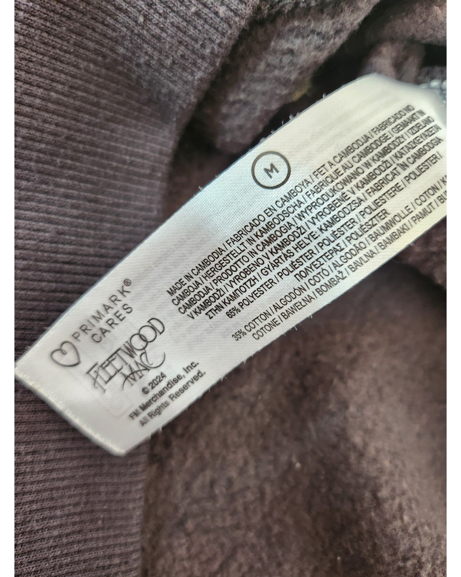 A close-up of a Primark label, size M, with care instructions and fabric details in several languages, attached to an upcycled grey Fleetwood Mac embroidered breastfeeding & pumping sweatshirt.