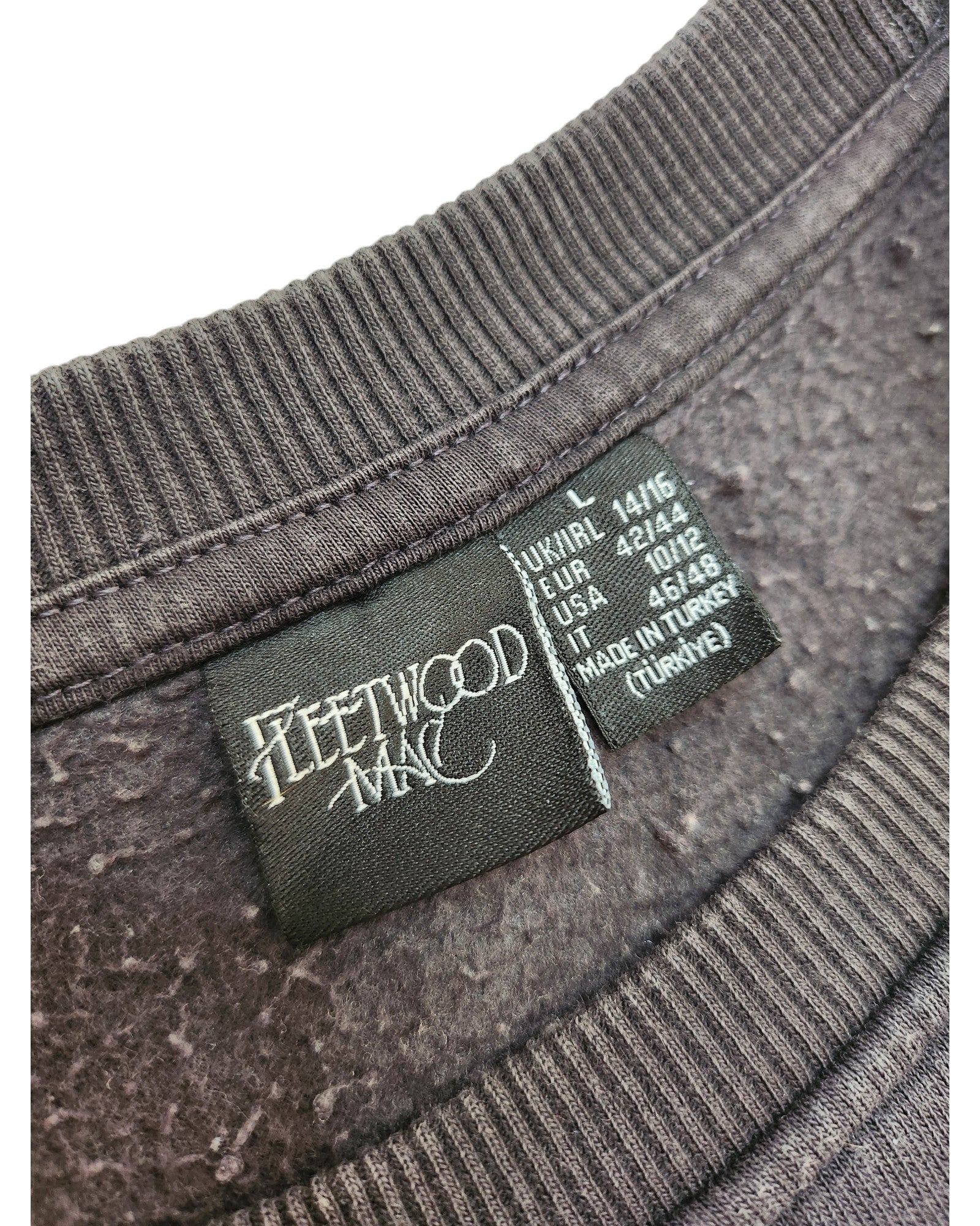 Close-up of a black label inside a dark grey Primark Upcycled Breastfeeding & Pumping Fleetwood Mac Floral Sweatshirt (L/UK14/16), showing the band’s logo, size info for various regions, and Made in Turkey at the bottom.