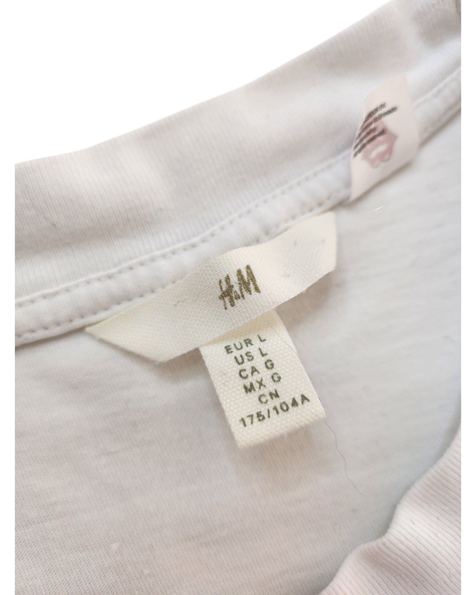 A close-up of an H&M tag showing sizes (EUR L, US L, CA G, MX G, CN 175/104A) on the neckline of the Upcycled Breastfeeding & Pumping White/Red Rolling Stones Band t-shirt (L), made from upcycled clothing.