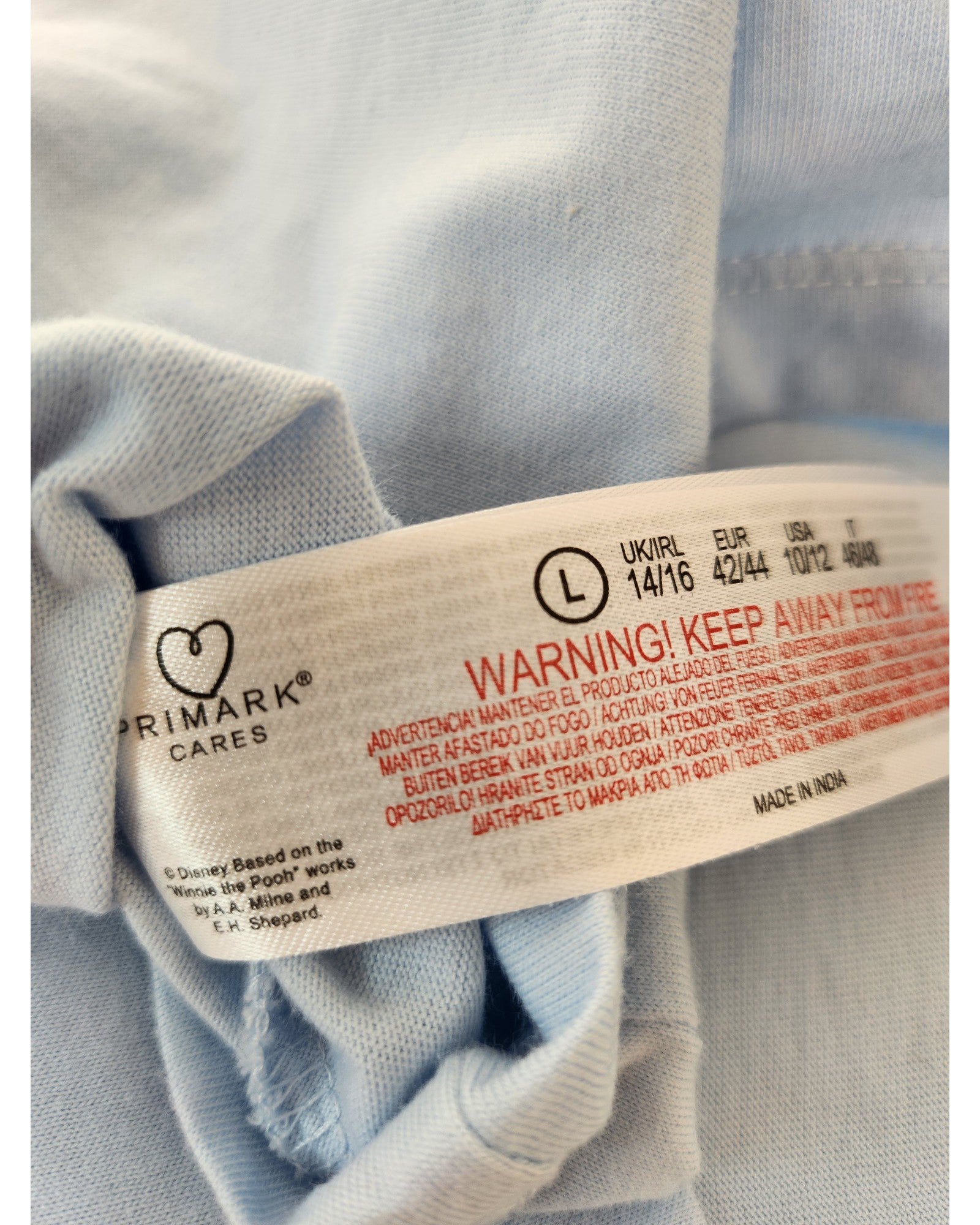A close-up of a Primark label on light blue Upcycled Breastfeeding & Pumping Piglet Disney Nightshirt (UK14/16) shows size, care instructions, and a red fire warning—highlighting eco-friendly fashion for mindful shoppers.