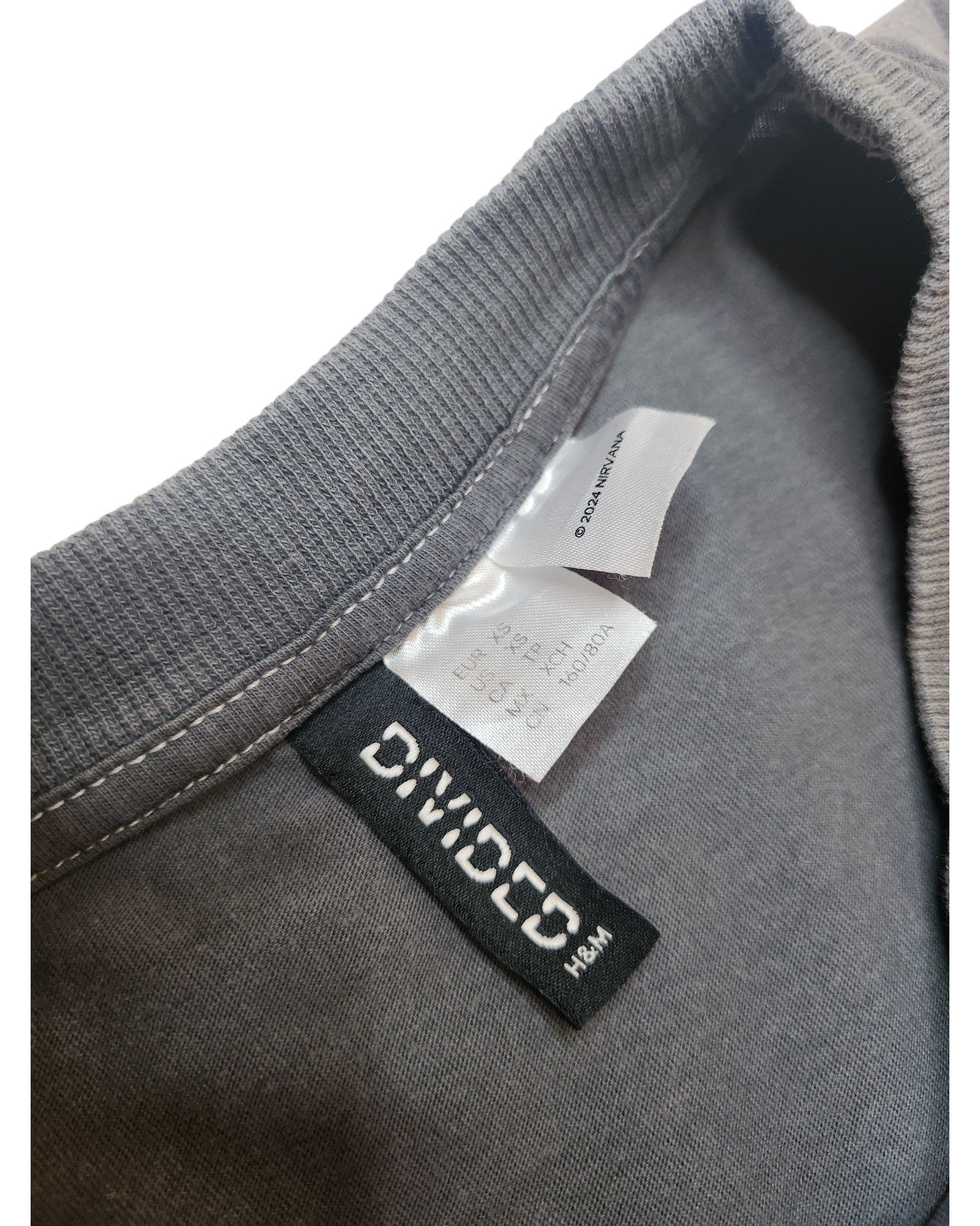 Close-up of a gray Upcycled Breastfeeding & Pumping Nirvana Guitar Band t-shirt by H&M (XS) shows the collar with tags, including DIVIDED H&M and care info—highlighting this unique preloved item.