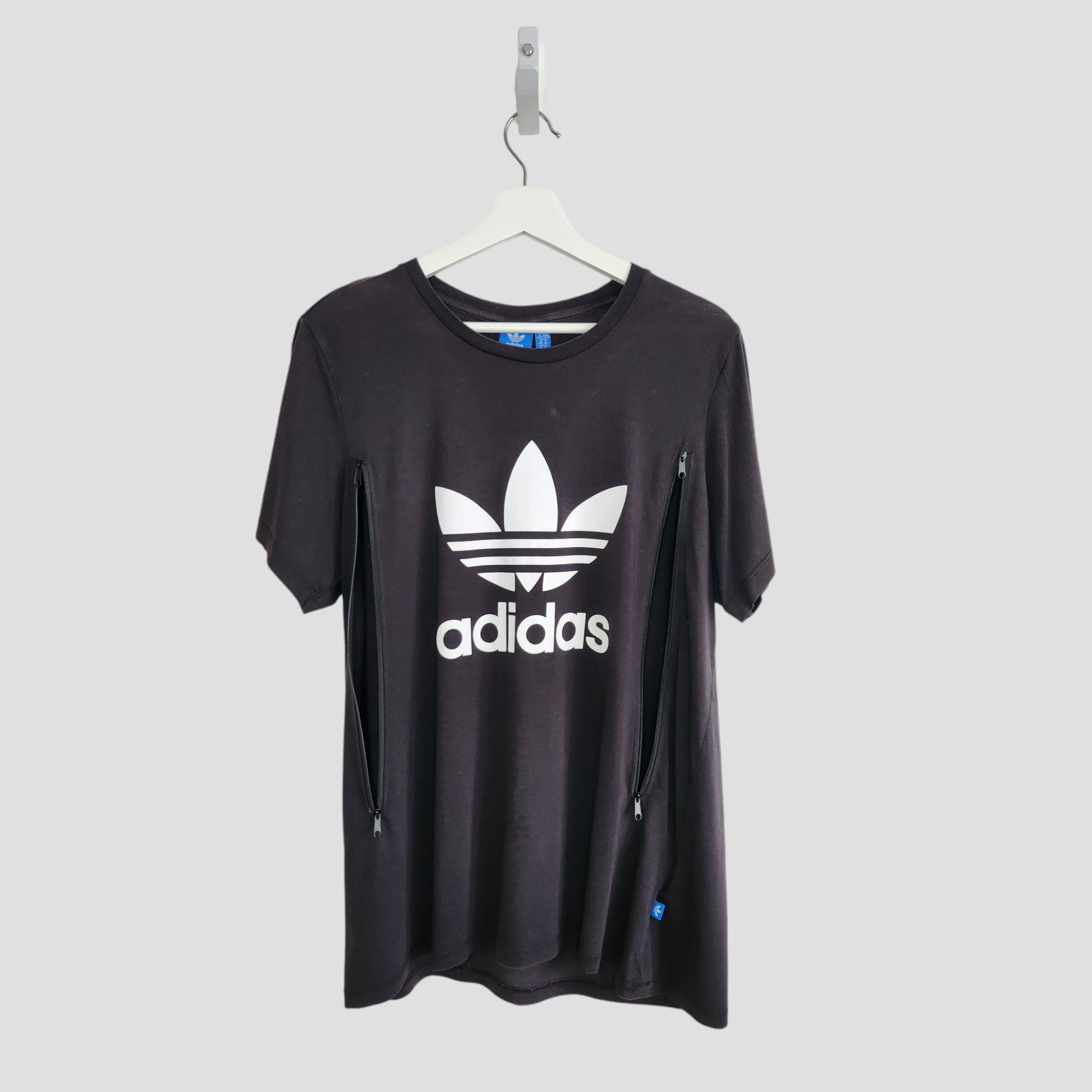 A preloved black/white Adidas UK14 t-shirt for breastfeeding & pumping, featuring a large white trefoil logo and text on the front, with side zippers for added style and convenience. Displayed on a white hanger against a light gray background.