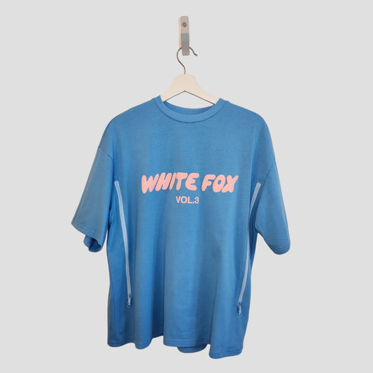 Upcycled breastfeeding & pumping blue White Fox t-shirt (M, 100265) with WHITE FOX VOL.3 in pink and white bubble letters on the back, featuring side zippers for nursing access. Displayed hanging against a light background. Brand: Unknown.
