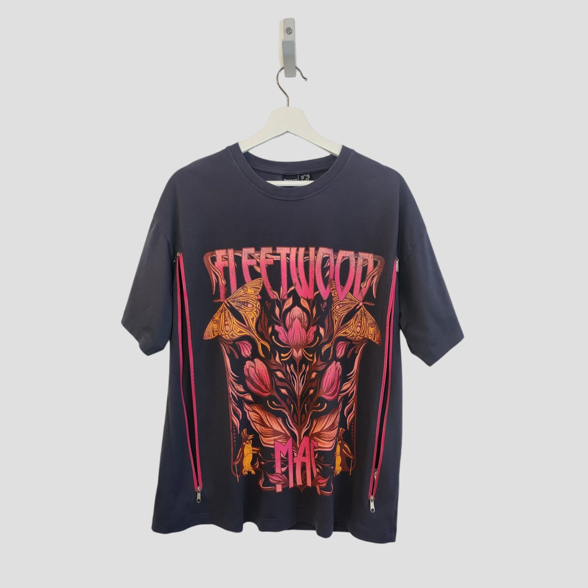 A grey floral Fleetwood Mac band t-shirt by Primark hangs on a white hanger. Upcycled for breastfeeding and pumping, it features bold pink/orange text, vibrant floral/moth graphics, and pink side zippers. Size S (UK10/12).
