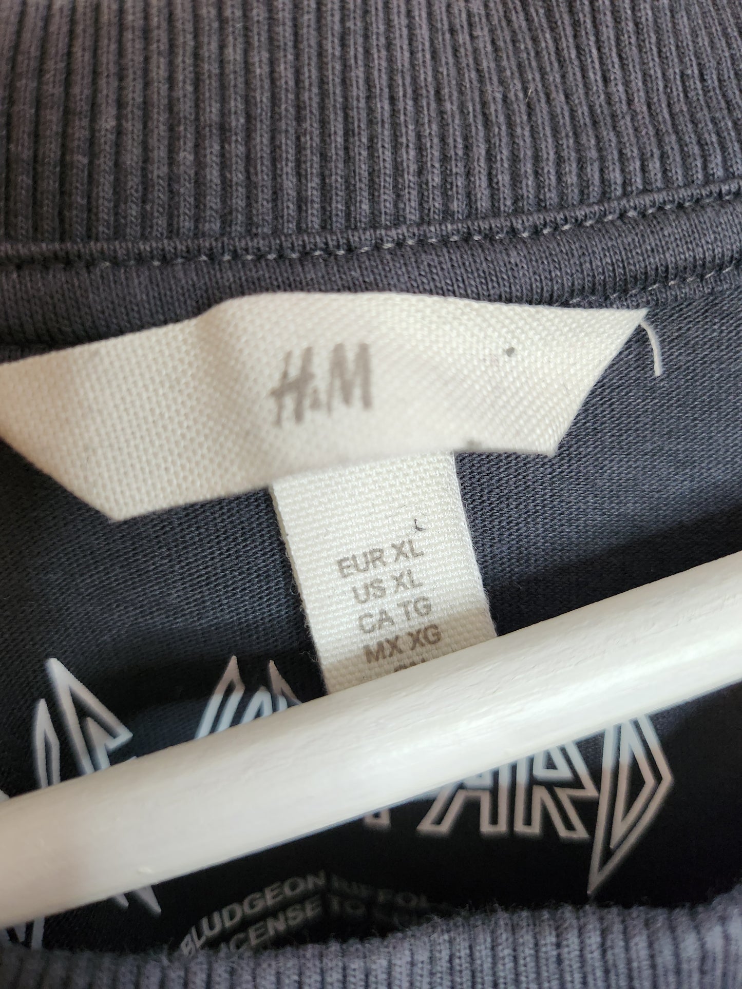 Close-up of an H&M label on an XL upcycled Def Leppard nursing & pumping t-shirt (100257) attached to dark ribbed fabric, with part of a white hanger and band logo visible—stylish preloved maternity wear.