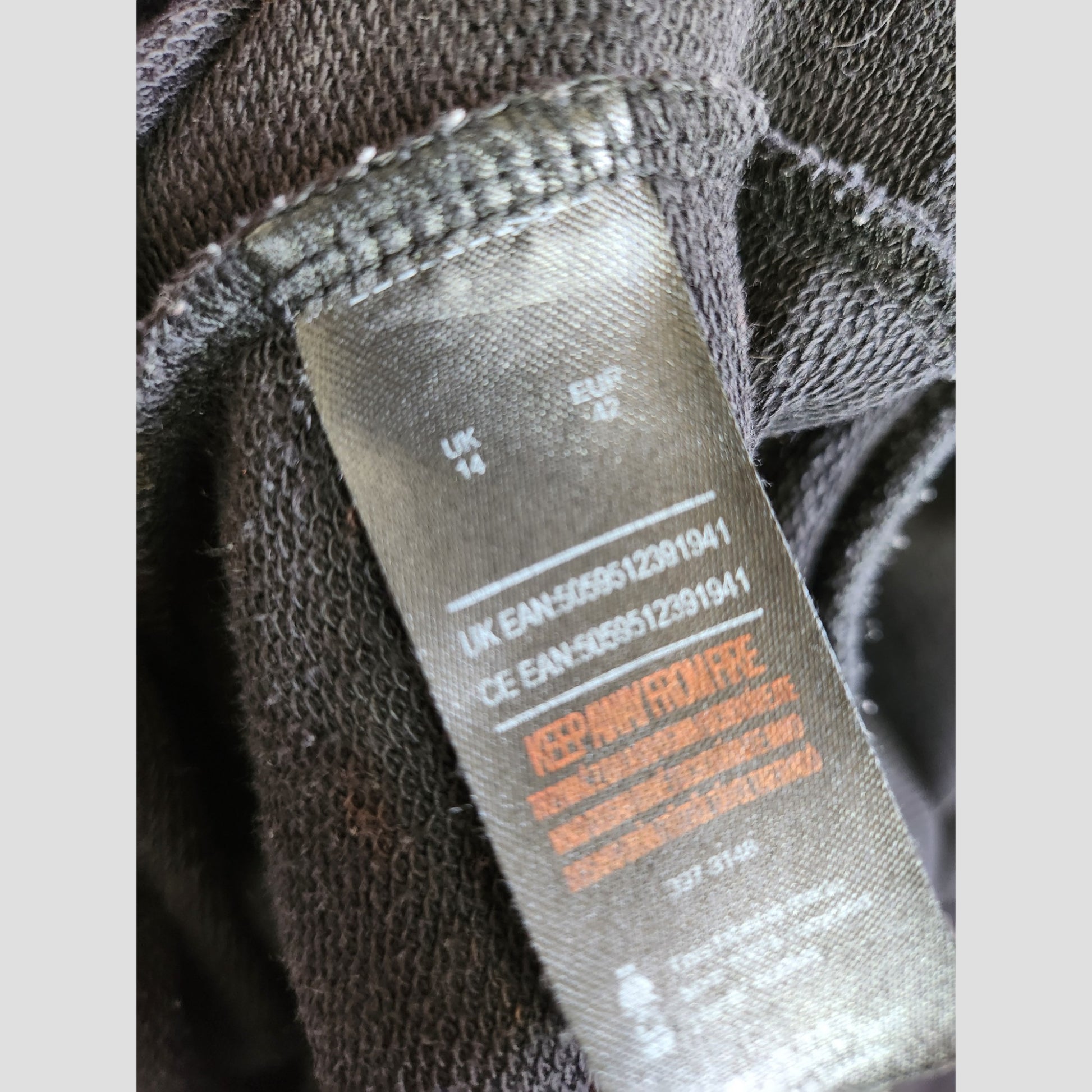 A close-up of the black clothing label on The Rolling Stones’ Upcycled Breastfeeding & Pumping Black/Leopard Band Sweatshirt UK14 (100280), displaying UK/EU sizes, barcode, and care info amid dark fabric from affordable eco fashion.