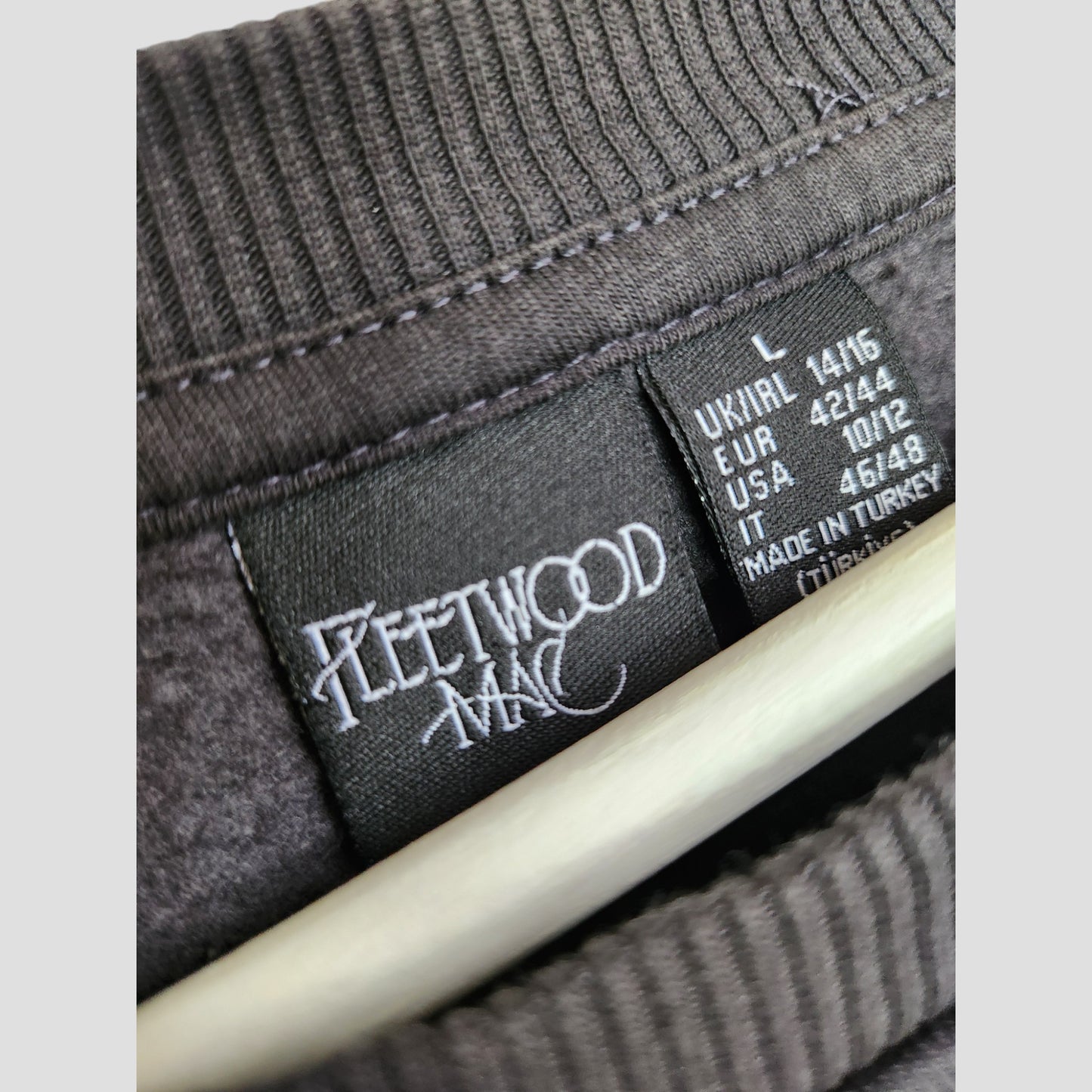 Close-up of a Primark clothing tag inside a dark grey Fleetwood Mac floral band sweatshirt (L, UK14/16), shown on a hanger—an example of upcycled breastfeeding & pumping eco-friendly nursing wear.