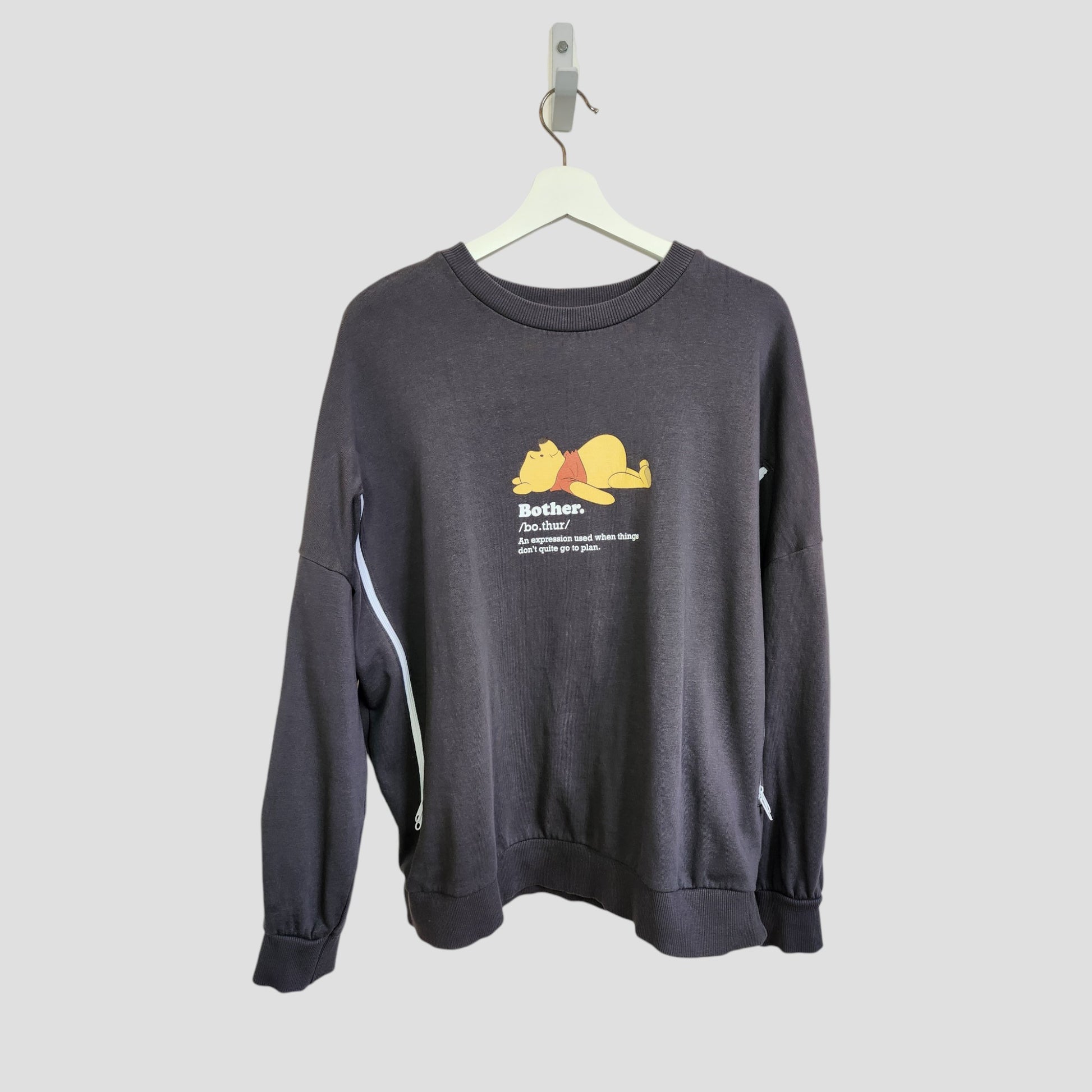 The George Upcycled Breastfeeding & Pumping Pooh Bother Sweatshirt (L, 100126) is a dark gray eco-friendly option featuring Winnie the Pooh lying down with “Bother.” and a quote on the front, displayed on a white hanger against a light gray background.