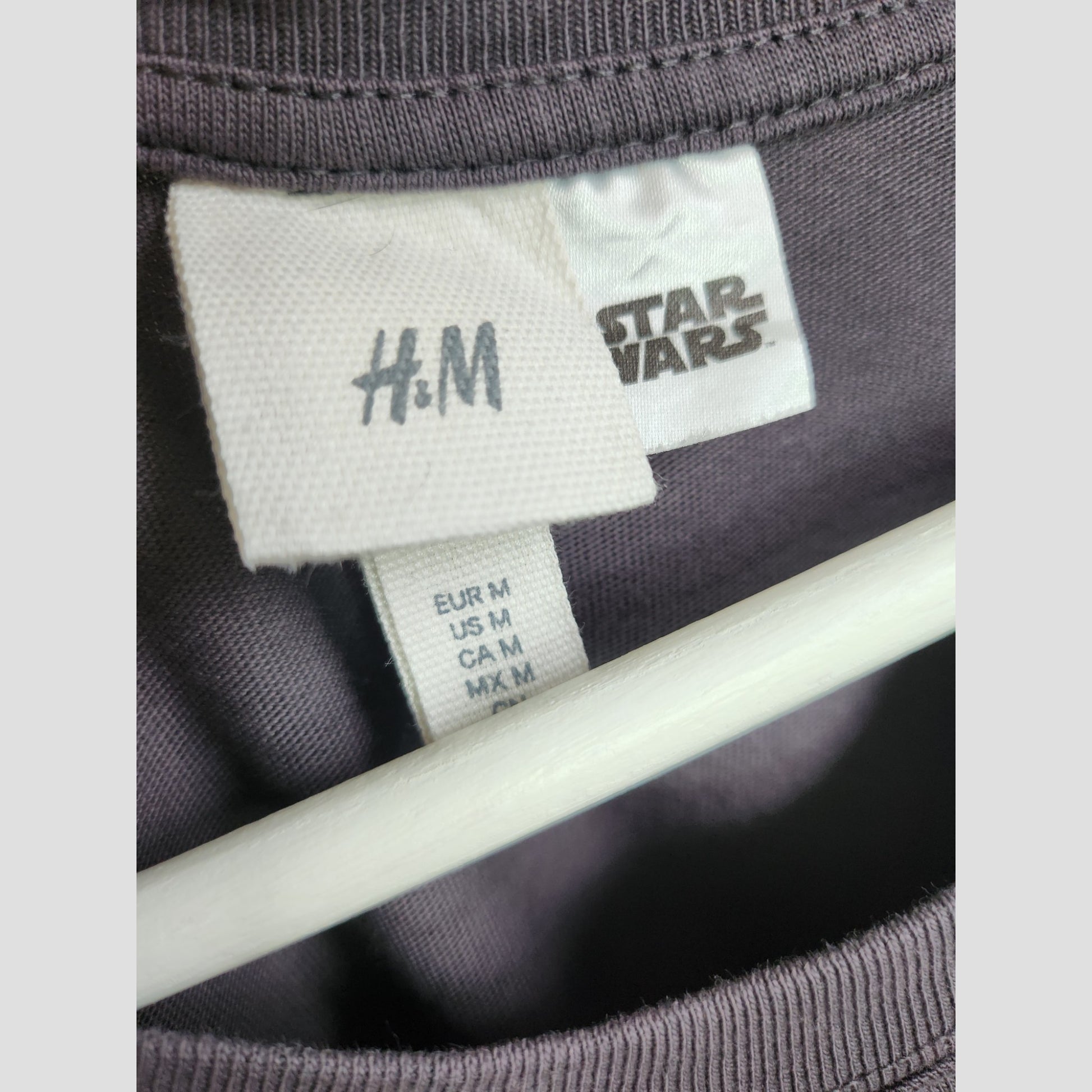 Close-up of H&M Upcycled Breastfeeding & Pumping Grey Star Wars t-shirt (M) tags, highlighting medium size for Europe, US, Canada, and Mexico. Eco-friendly item on a white hanger.