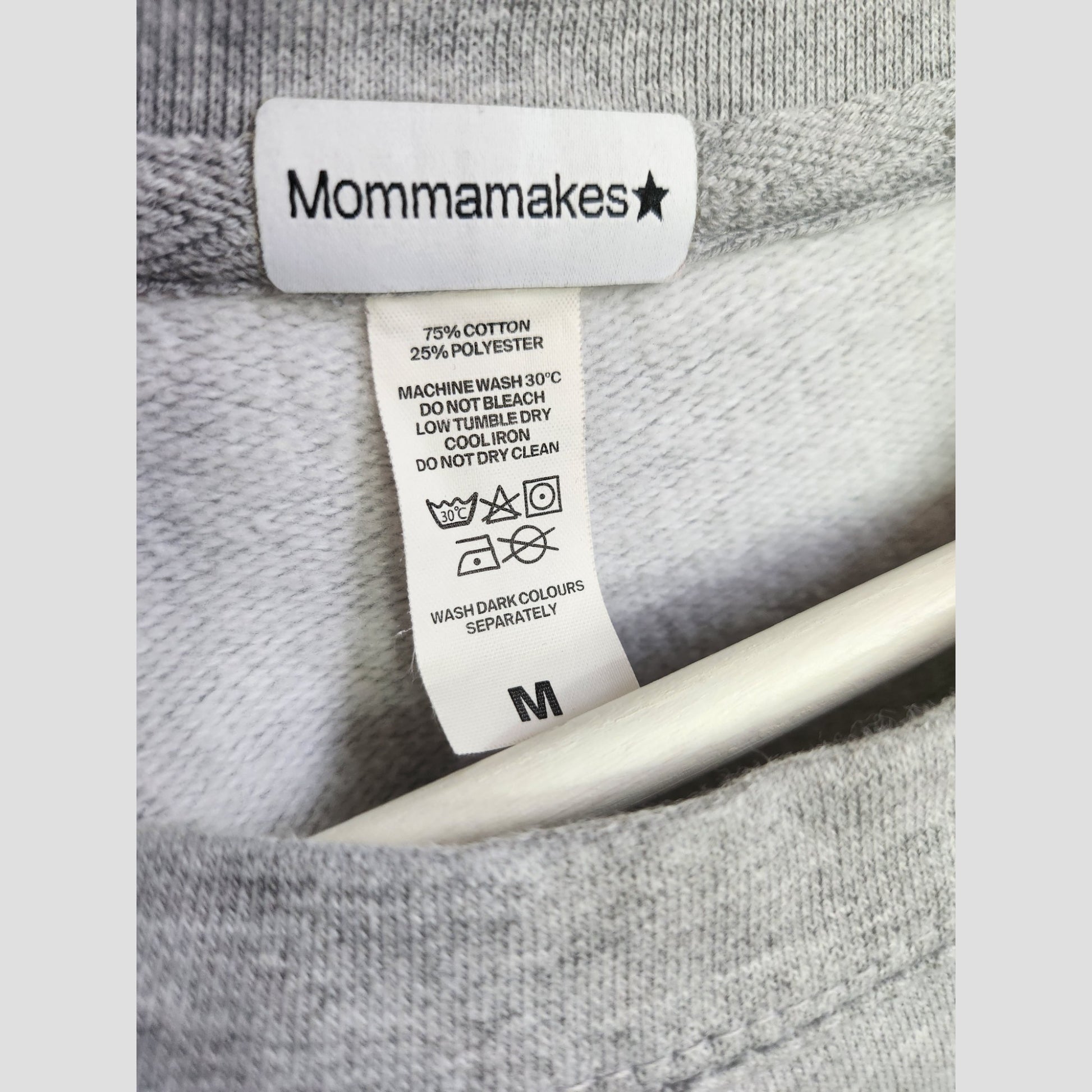 Close-up of a gray Upcycled Breastfeeding & Pumping Tired Mums Club Sweatshirt (M) label by Momma Makes, showing size “M” and eco-conscious care instructions for washing, ironing, and bleaching.