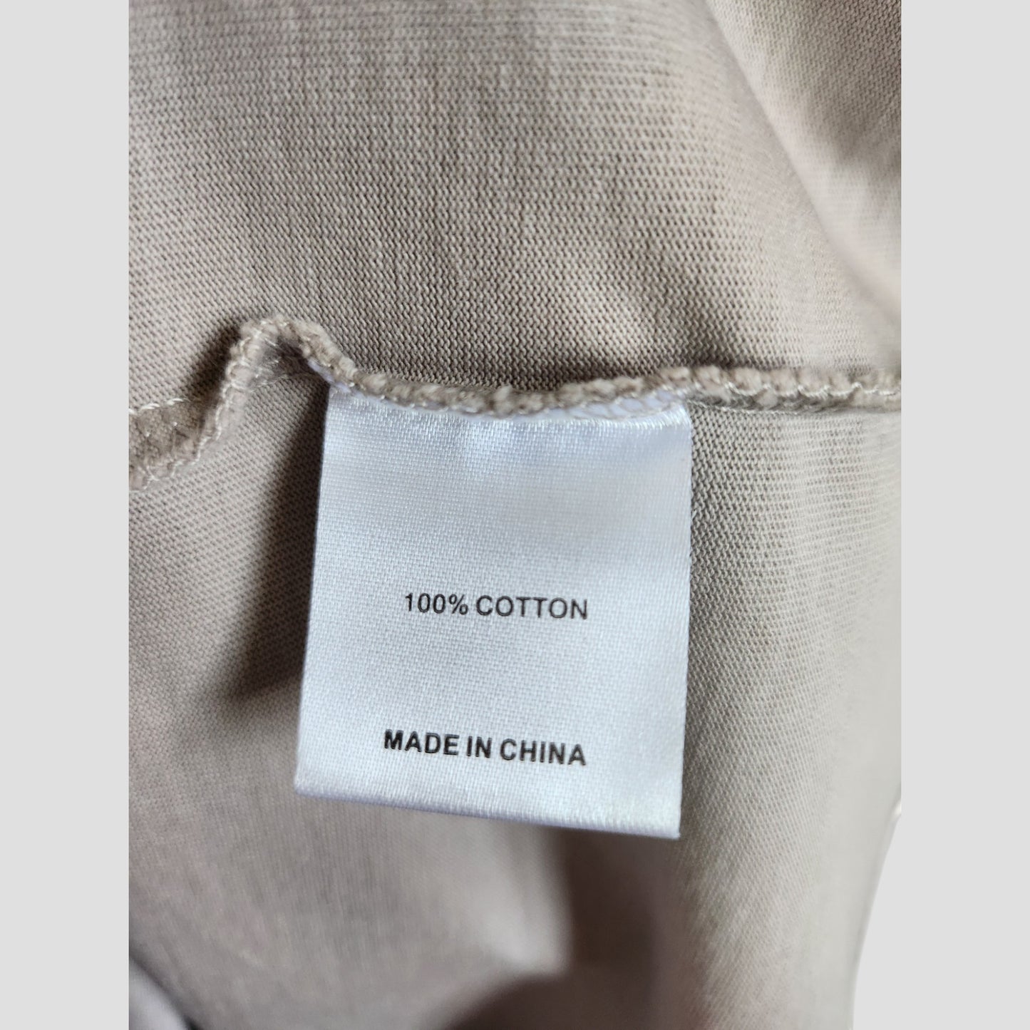 A close-up of a white clothing tag on beige fabric for the Upcycled Breastfeeding & Pumping Grey/Light Blue White Fox t-shirt (L, 100307), showing "100% COTTON" and "MADE IN CHINA" in black text. Brand: Unknown.