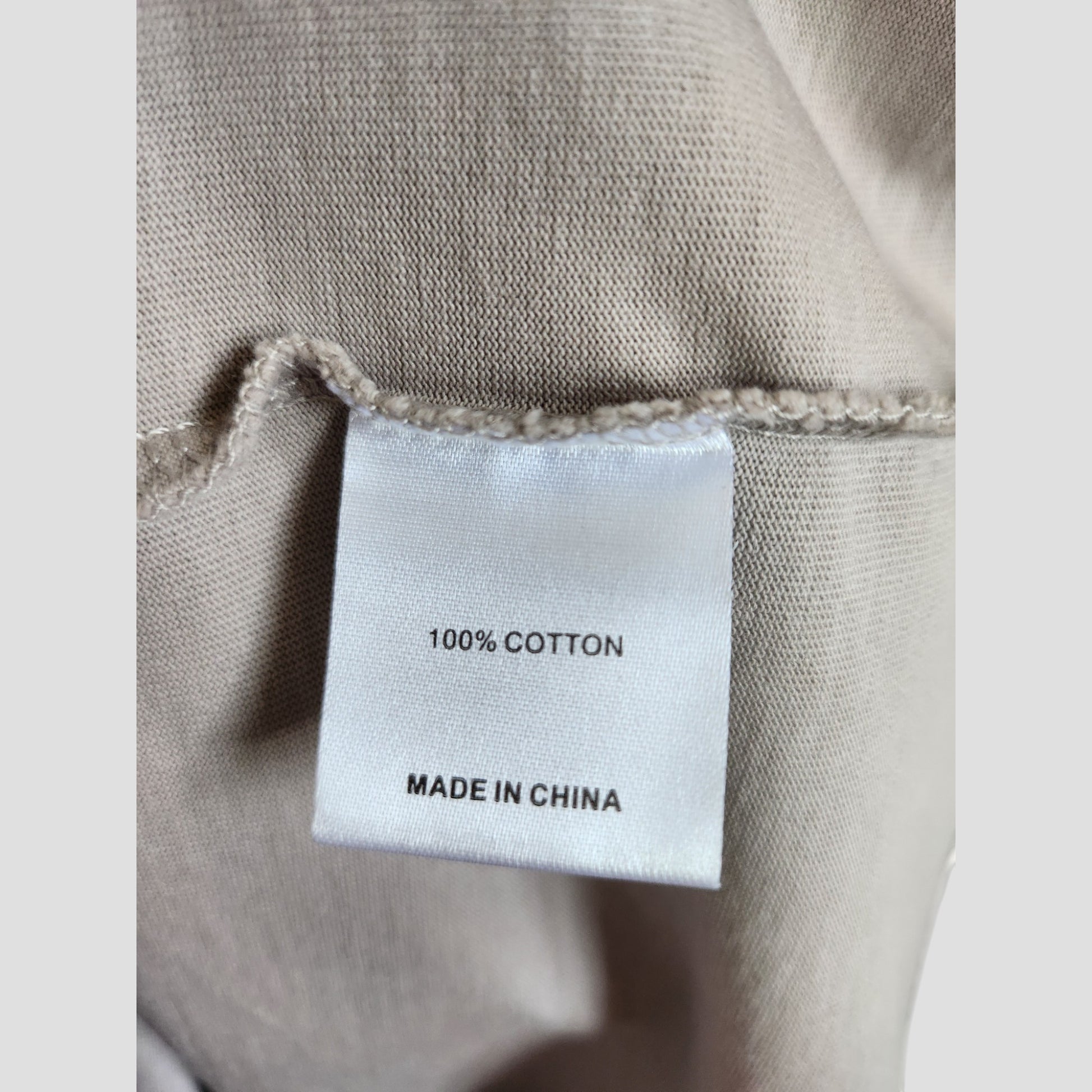 A close-up of a white clothing tag on beige fabric for the Upcycled Breastfeeding & Pumping Grey/Light Blue White Fox t-shirt (L, 100307), showing "100% COTTON" and "MADE IN CHINA" in black text. Brand: Unknown.