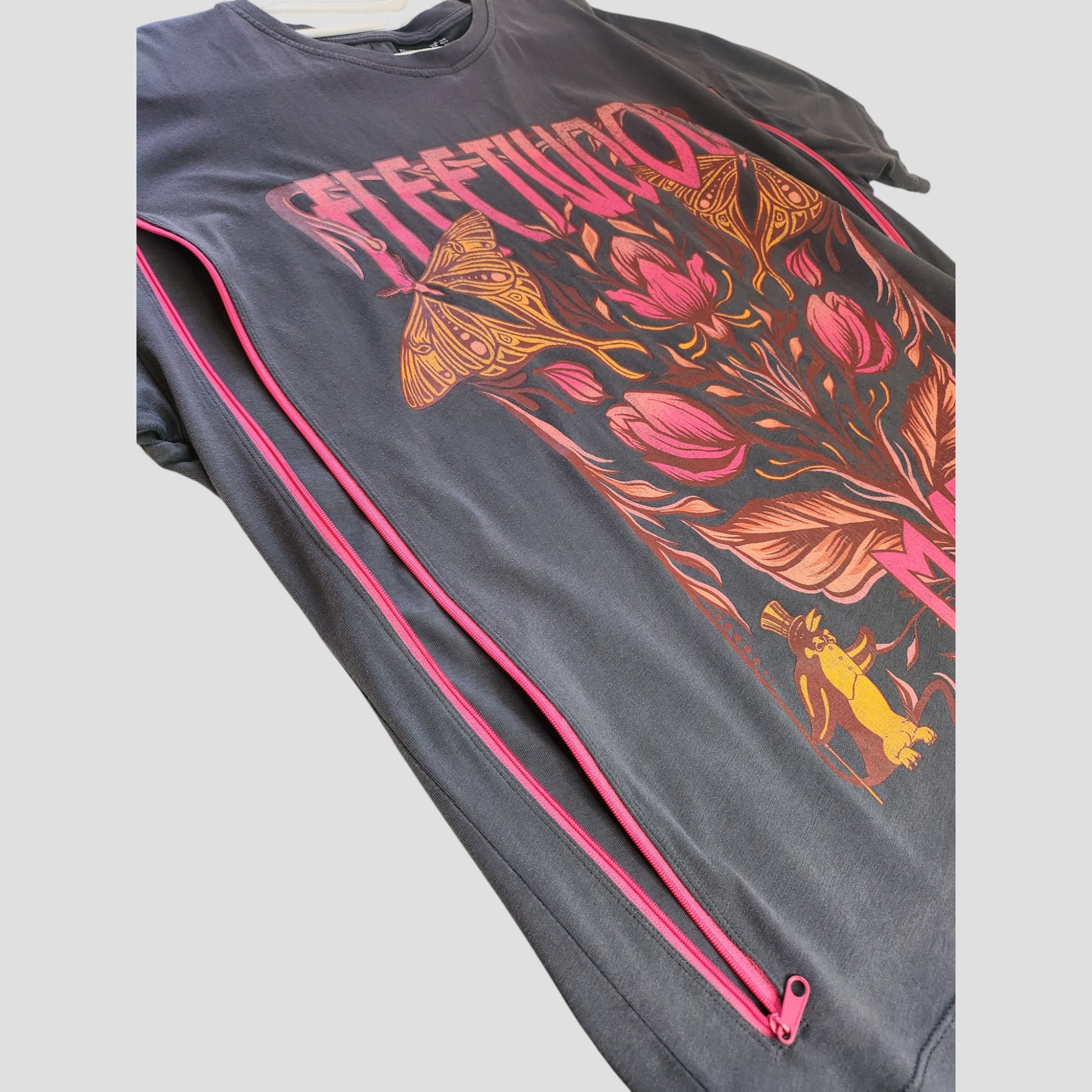 A close-up of Primark’s upcycled dark grey Fleetwood Mac floral band nursing t-shirt (L, UK14/16), featuring flower, moth, and cat graphics, bold text, and a pink diagonal zipper—ideal for breastfeeding or pumping fans.