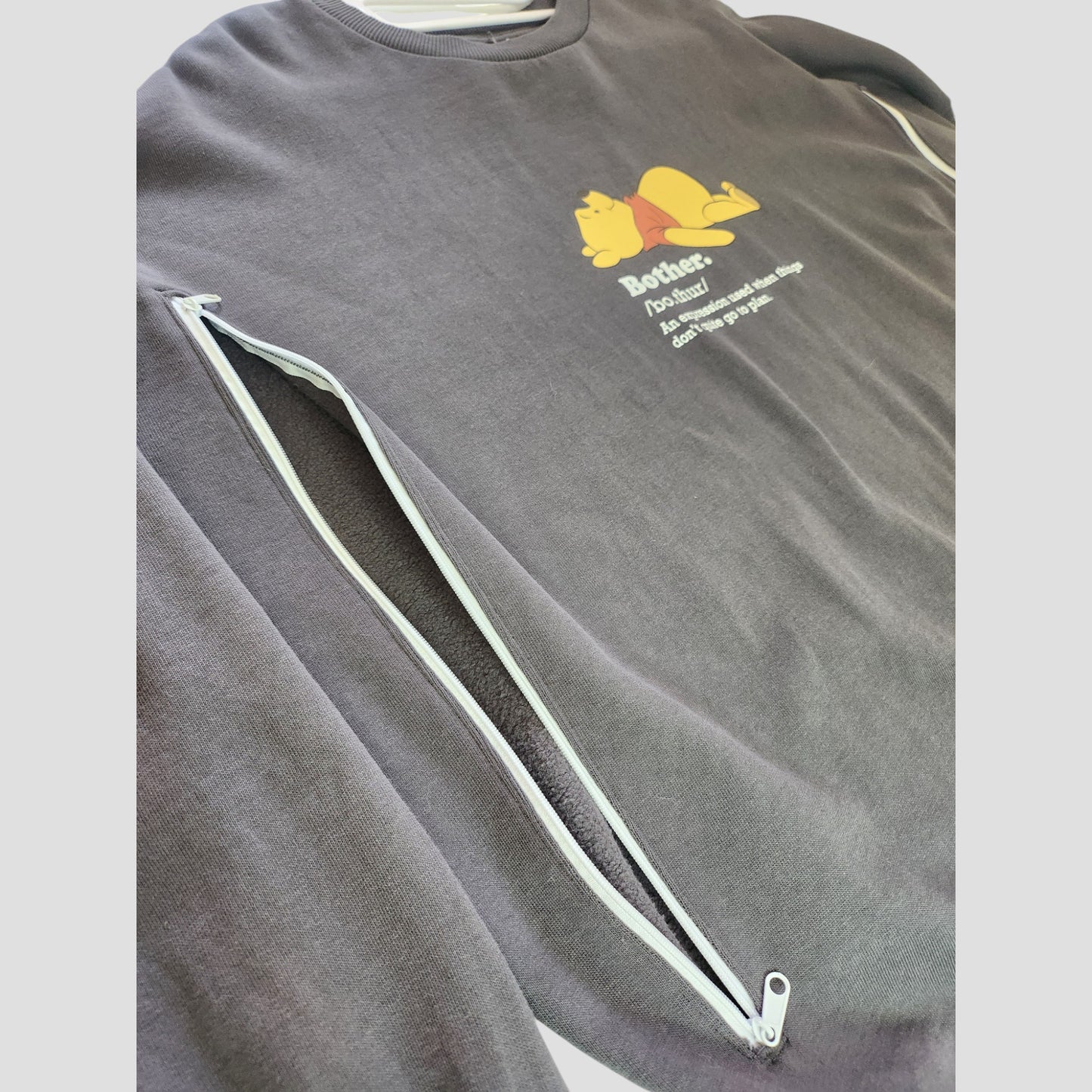The George Upcycled Breastfeeding & Pumping Pooh Bother Sweatshirt (L, 100126) features a gray design with horizontal white zippers and a yellow cartoon cat. Made from upcycled materials for comfort—perfect for nursing or napping.
