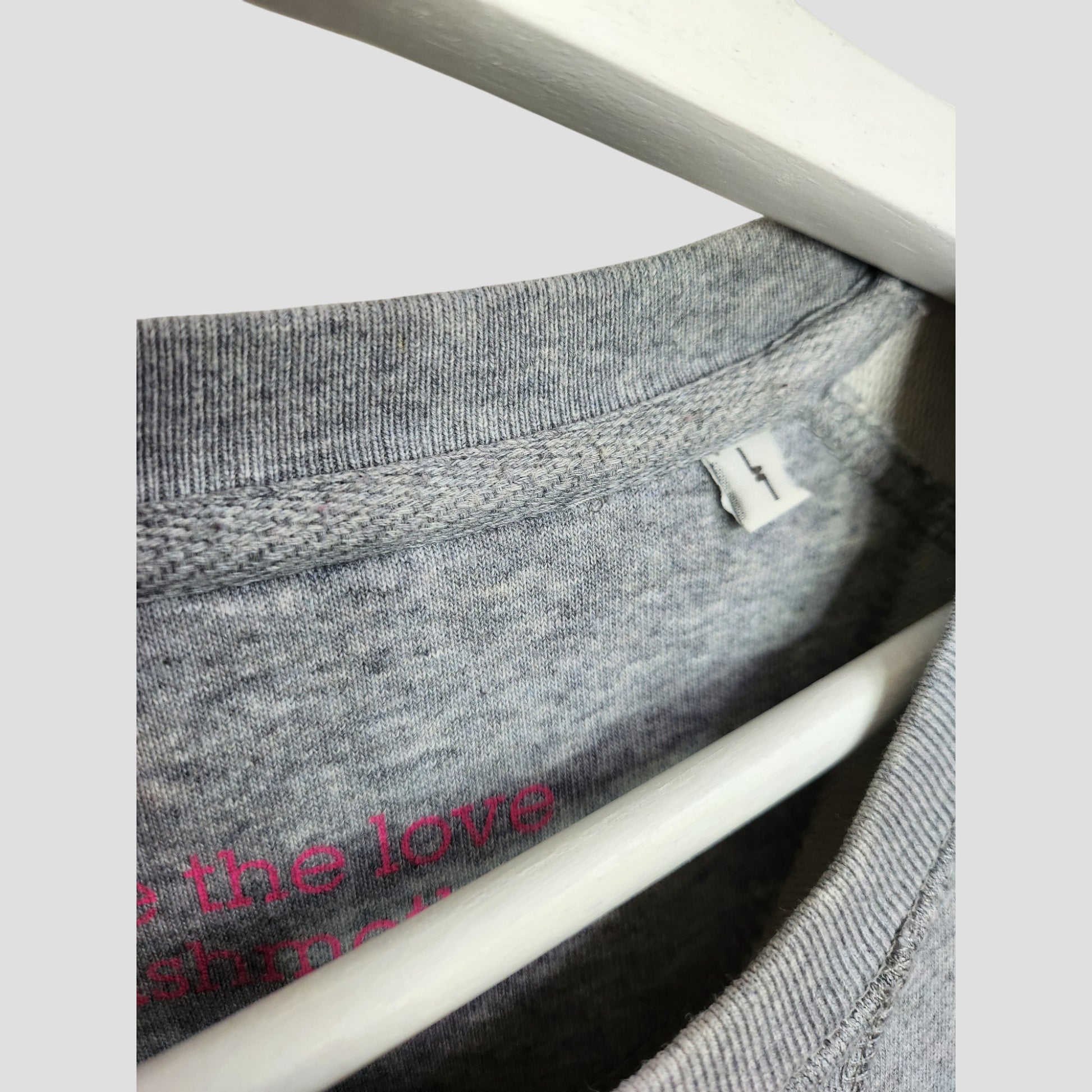A close-up of the Selfish Mother Upcycled Breastfeeding & Pumping Grey/Pink MOTHER Sweatshirt (L) on a white hanger shows the inside collar with a white size L tag and part of pink printed text on the eco-friendly fabric.