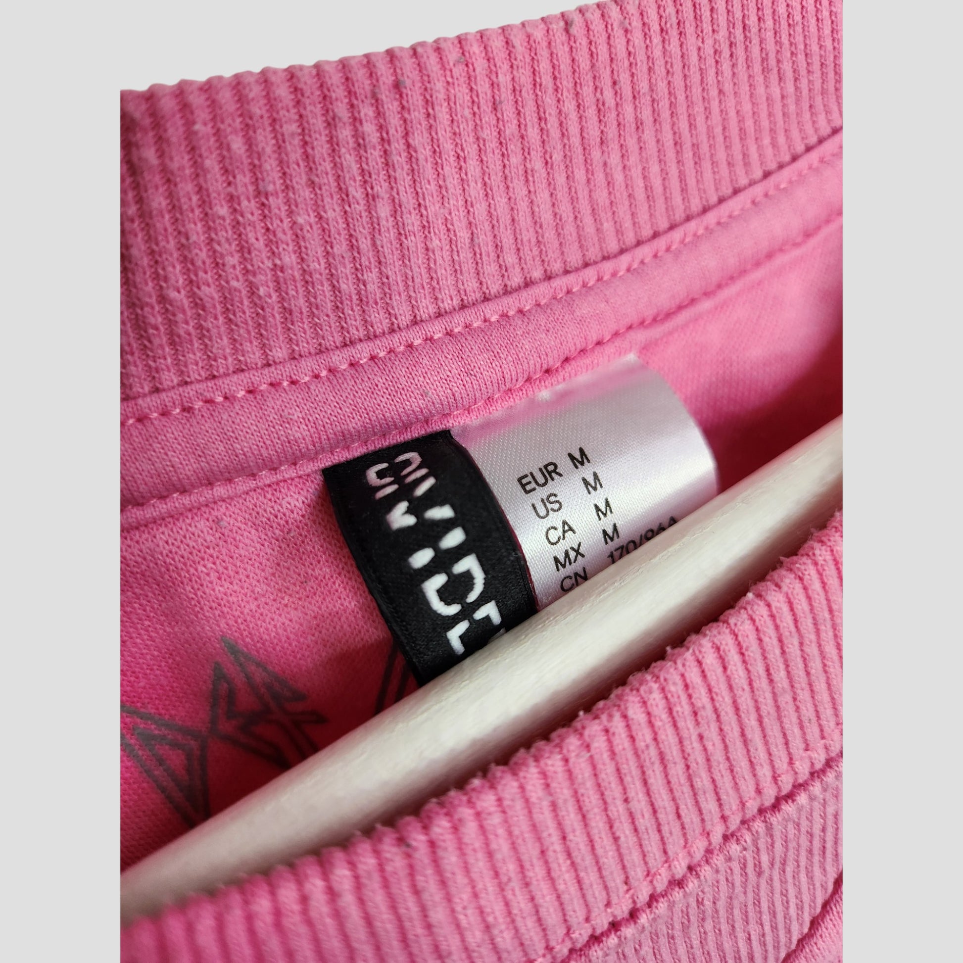 Close-up of a pink H&M Upcycled Breastfeeding & Pumping Def Leppard Band Sweatshirt (M, 100131) collar with regional size tag and brand label, emphasizing its eco-friendly, pre-loved origins. A hanger is visible beneath.