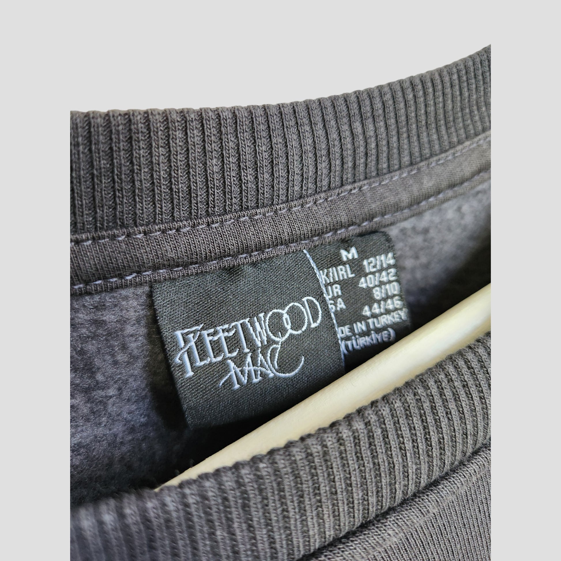 A close-up of a Primark Upcycled Breastfeeding & Pumping Dark Grey Fleetwood Mac Floral Band Oversized Sweatshirt’s collar and label, showing the logo, multi-language sizing info, and that this affordable eco piece is made in Turkey.