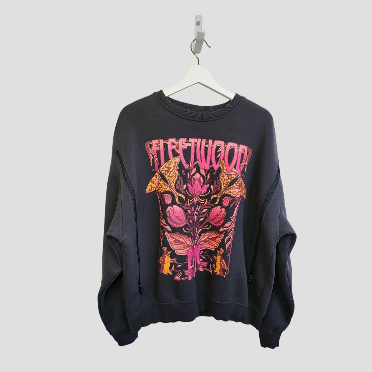A dark grey oversized Fleetwood Mac floral band sweatshirt by Primark (size M) hangs on a white hanger, crafted from upcycled breastfeeding clothing for eco-friendly style. Features psychedelic flowers, mushrooms, and the band name on the front.