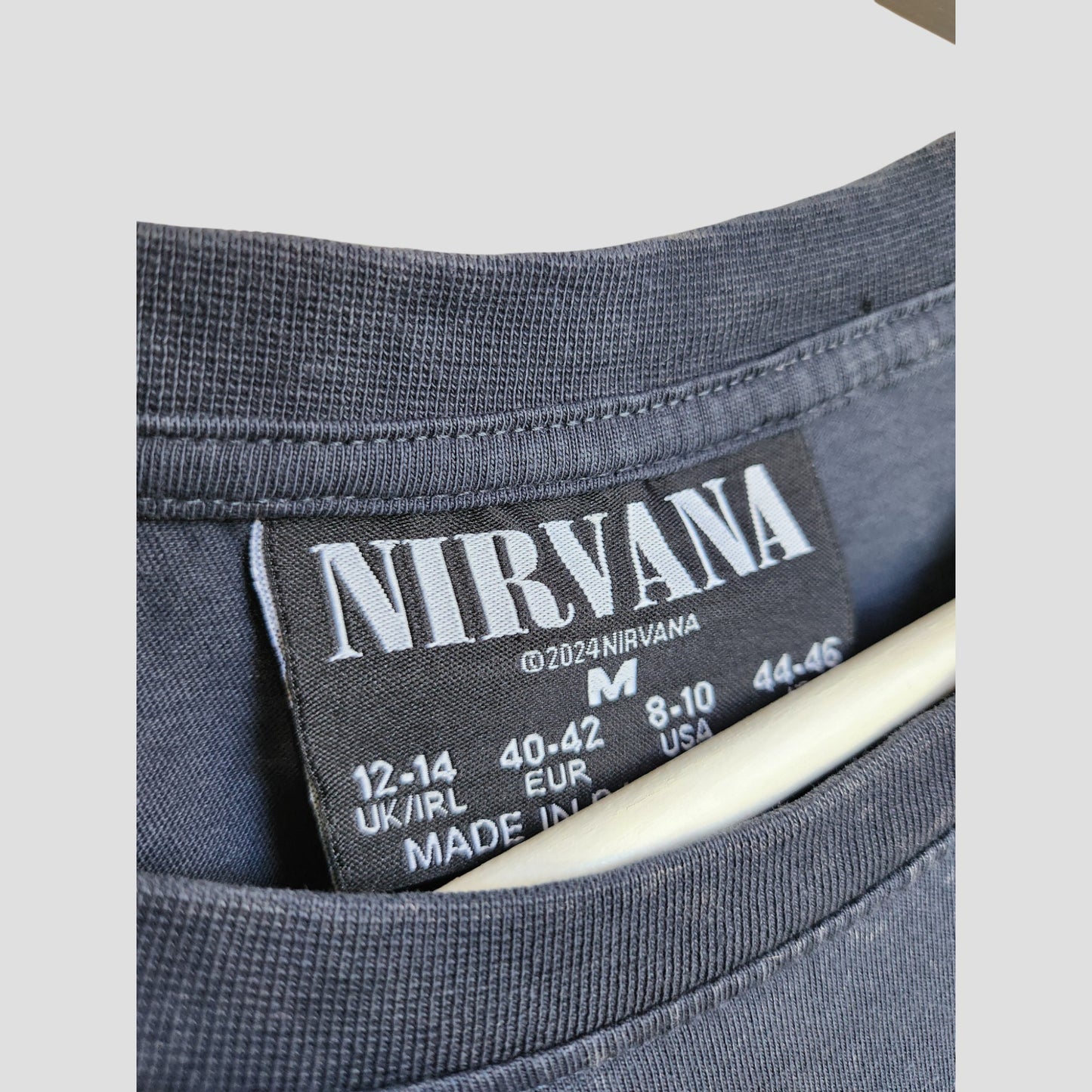 Close-up of a gray Primark t-shirt collar with a black label showing "NIRVANA" in white. This upcycled oversized breastfeeding & pumping Nirvana Angel Band tee (UK12/14, M) is perfect for music-loving moms; hanger visible under the collar.