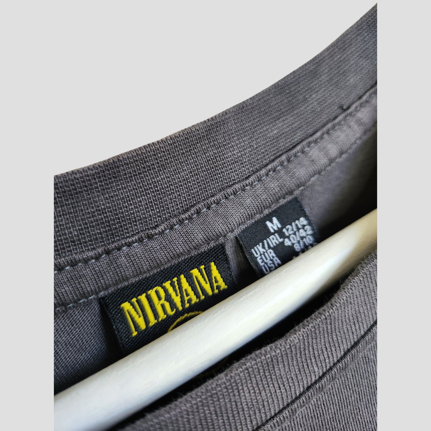 Close-up of a Primark Upcycled Breastfeeding & Pumping Grey/Yellow Smile Nirvana Band t-shirt (M/UK12/14, 100243) on a white hanger, featuring yellow Nirvana label and fabric details.