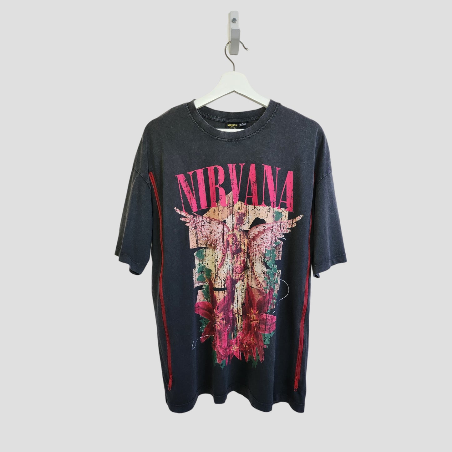 A dark grey floral Nirvana band oversized t-shirt by Primark hangs on a white hanger. This upcycled nursing and pumping tee (UK10/12) features a colorful angel and flower graphic with “NIRVANA” in bold pink letters across the chest.