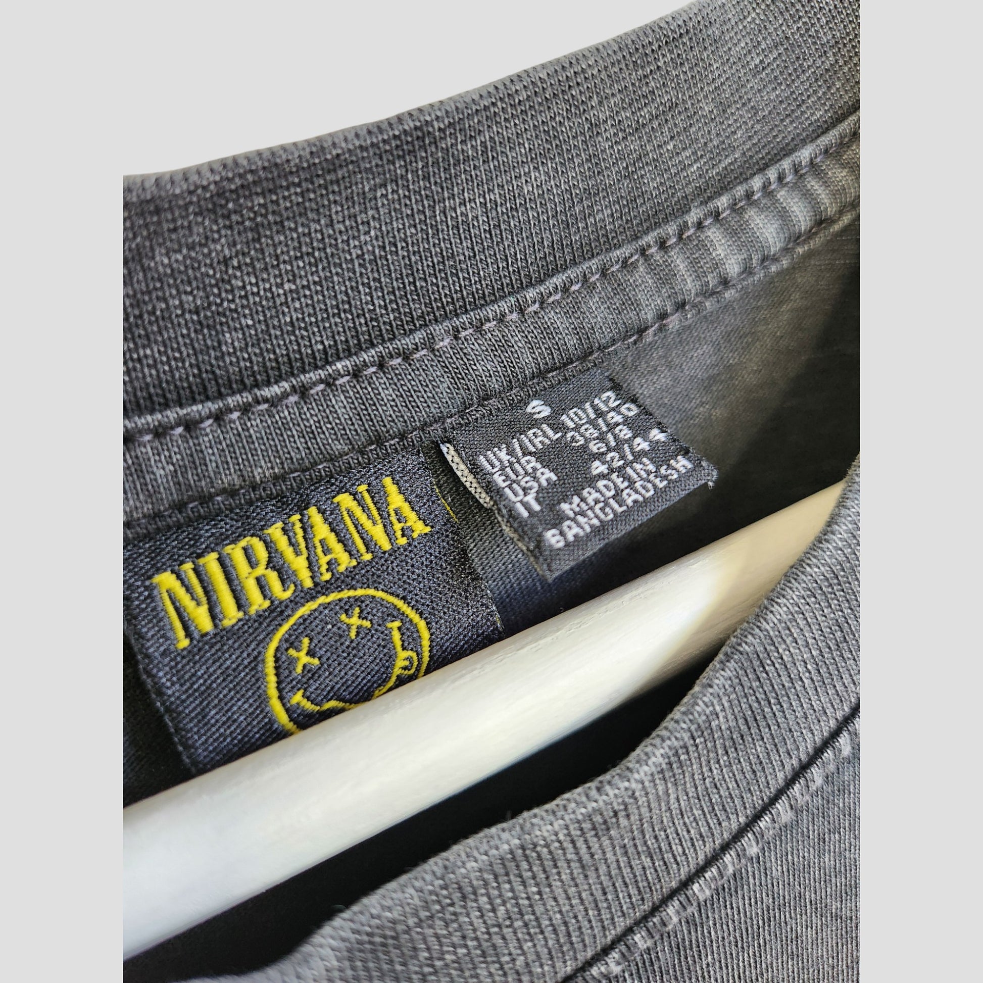 Close-up of a dark grey Primark Nirvana band T-shirt collar with floral design, yellow smiley label, and black care tag—an upcycled, eco-friendly oversized breastfeeding/pumping top in size S (UK10/12).