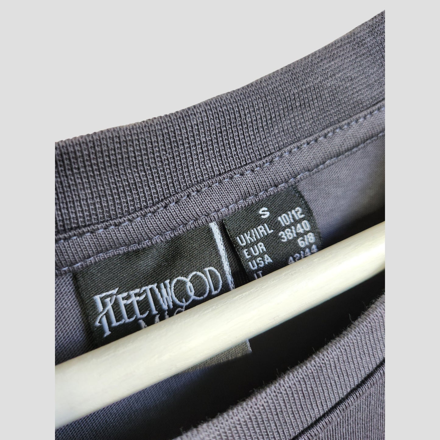 Close-up of a grey Primark t-shirt, featuring a "Fleetwood Mac Floral Band" design and size tag. This upcycled oversized nursing tee (UK 10/12) offers comfort and style for eco-conscious mums seeking preloved breastfeeding wear.