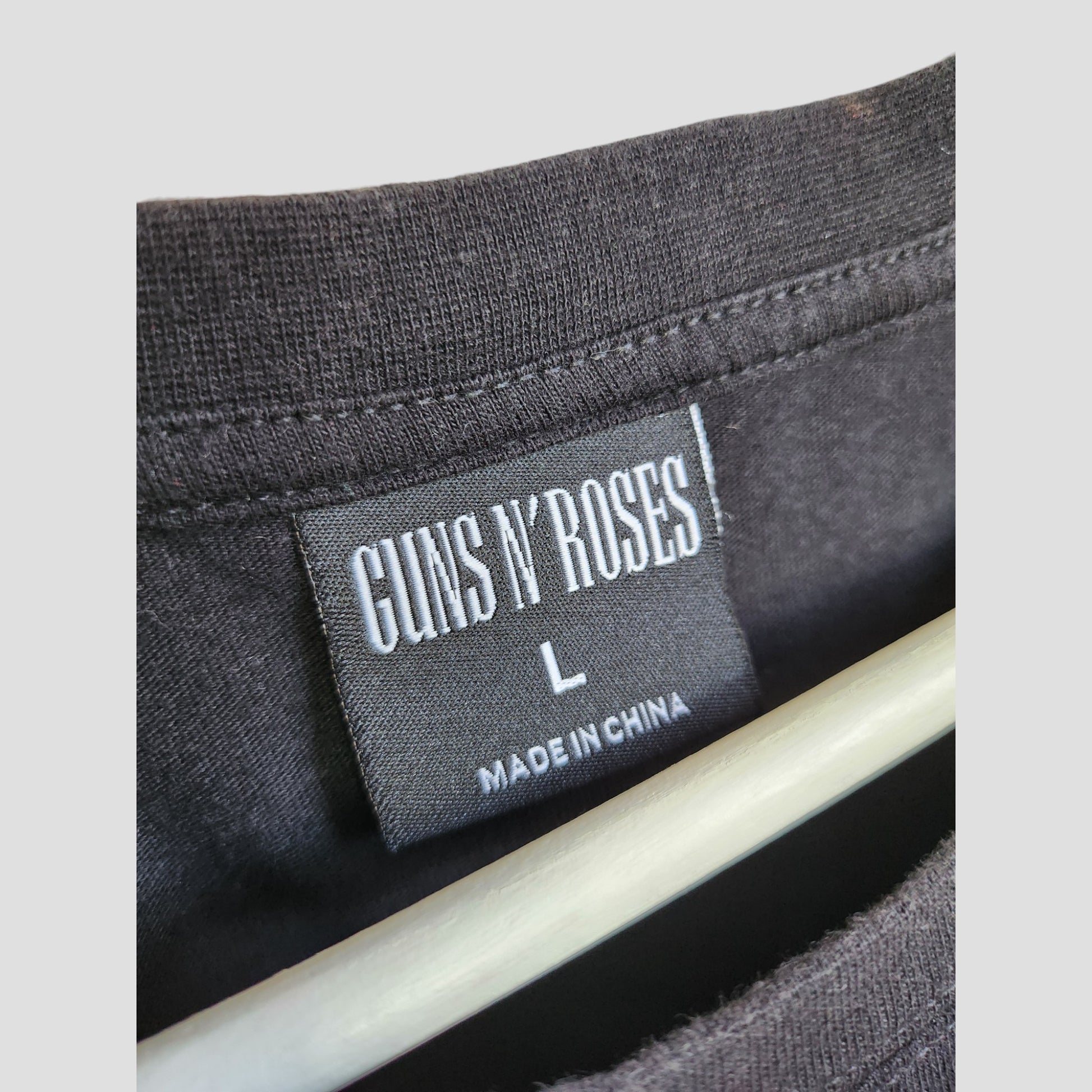 Close-up of a Primark upcycled breastfeeding & pumping black Guns N’ Roses band t-shirt (L, UK14/16) with a black tag reading "Guns N’ Roses L Made in China," hanging on a light-colored hanger.