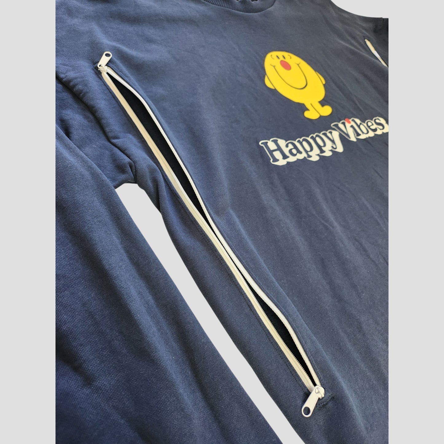 The Comic Relief Upcycled Breastfeeding & Pumping Happy Vibes Sweatshirt L (100127) is a blue top with "Happy Vibes" text and a smiling yellow character, featuring a white diagonal sleeve zipper for stylish, eco-friendly nursing access.