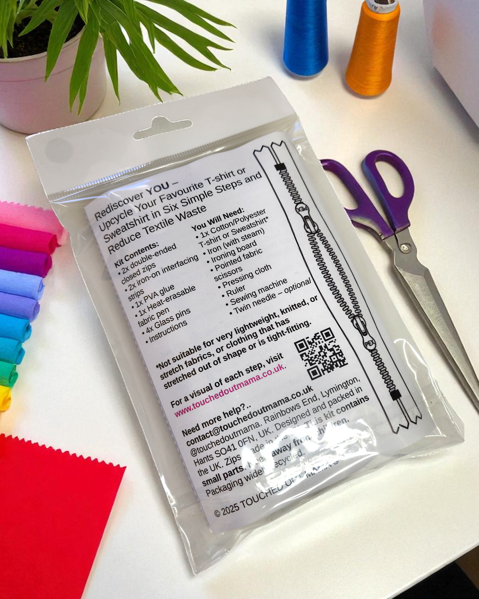 The Original Sew-On Zip Kit by touched out mama sits sealed on a white table with colorful thread spools, purple scissors, and multicolored fabric—ready to reduce textile waste. Green plant leaves peek in from the top left corner.