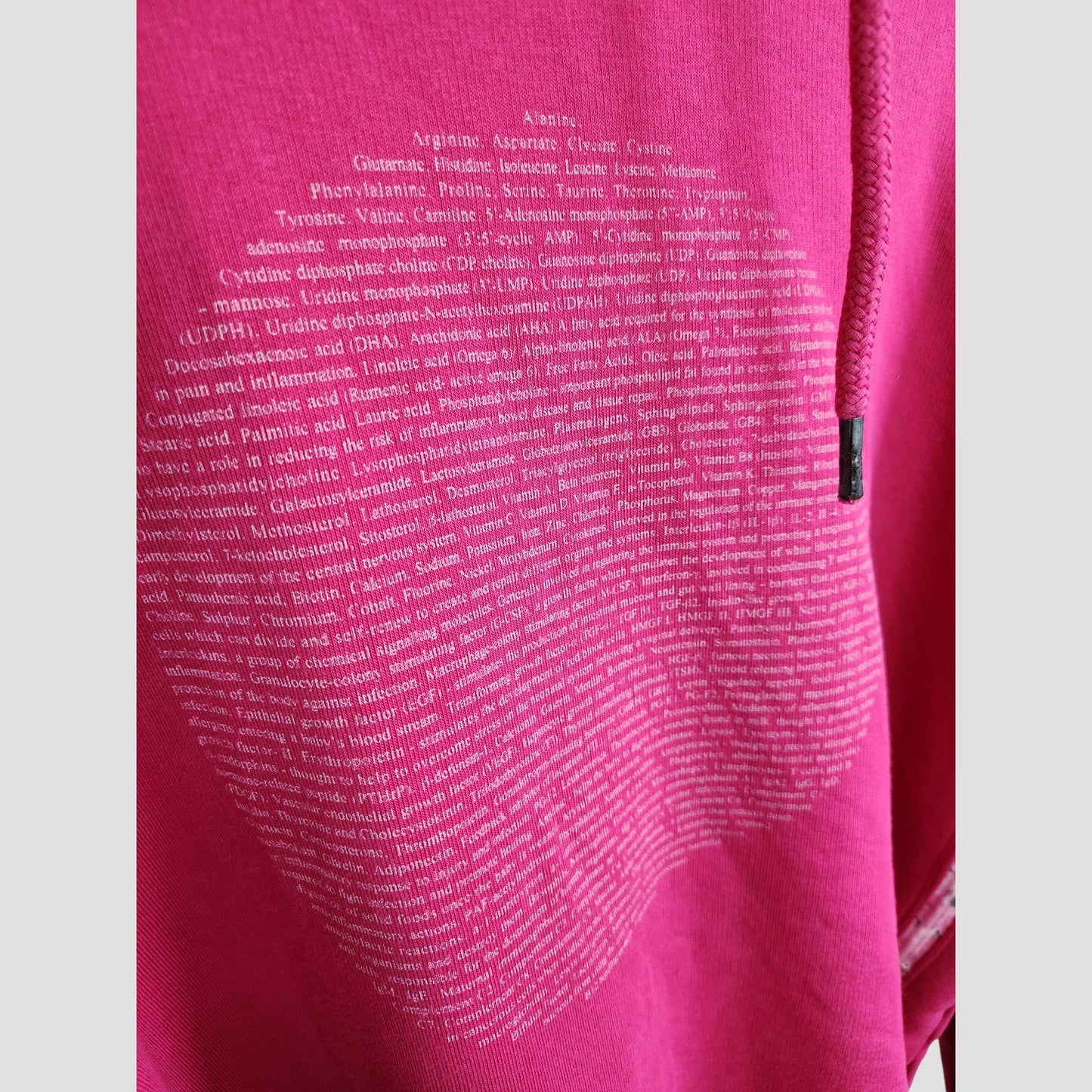 Close-up of the Human Milk Upcycled Breastfeeding & Pumping Human Milk Composition Hoodie L, showing pink fabric with white biochemical text in an apple shape and a metal-tipped drawstring visible on the right edge.