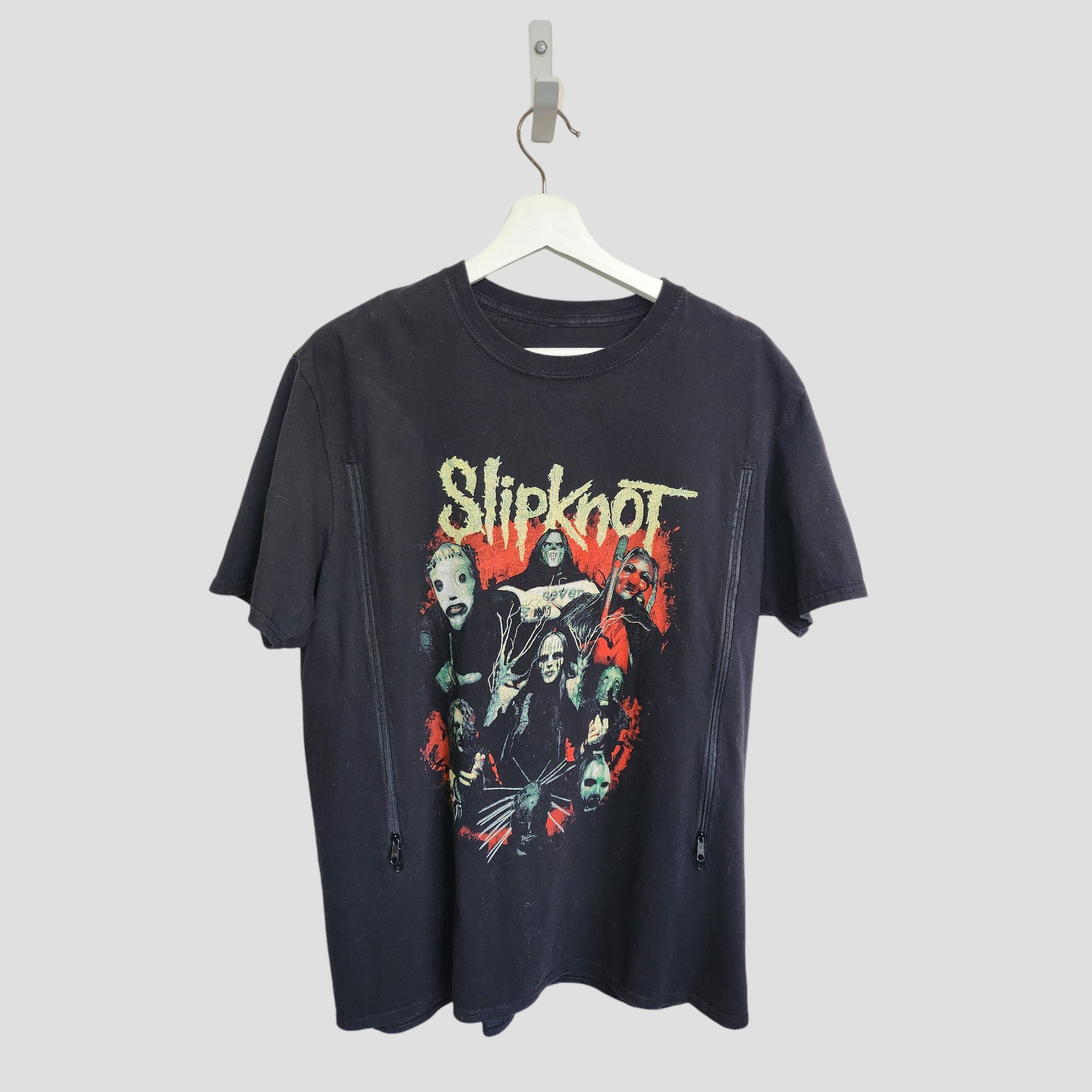 A preloved Slipknot upcycled breastfeeding & pumping t-shirt (L) in black hangs on a white hanger, showcasing masked band members in red/green tones with the band's distressed yellow logo above.
