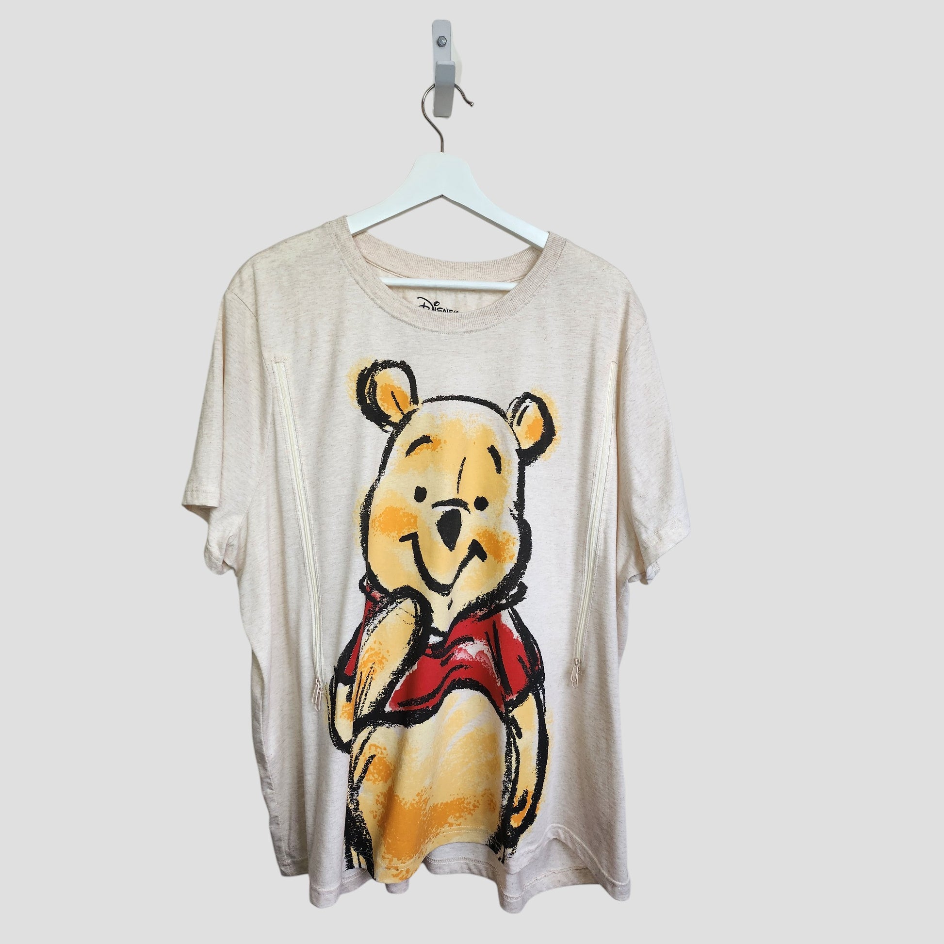 The Primark Upcycled Breastfeeding & Pumping Cream Winnie the Pooh T-shirt (2XL, UK22/24, 100364) features a large colorful graphic of Pooh in a red shirt and hangs on a white hanger against a plain light background.