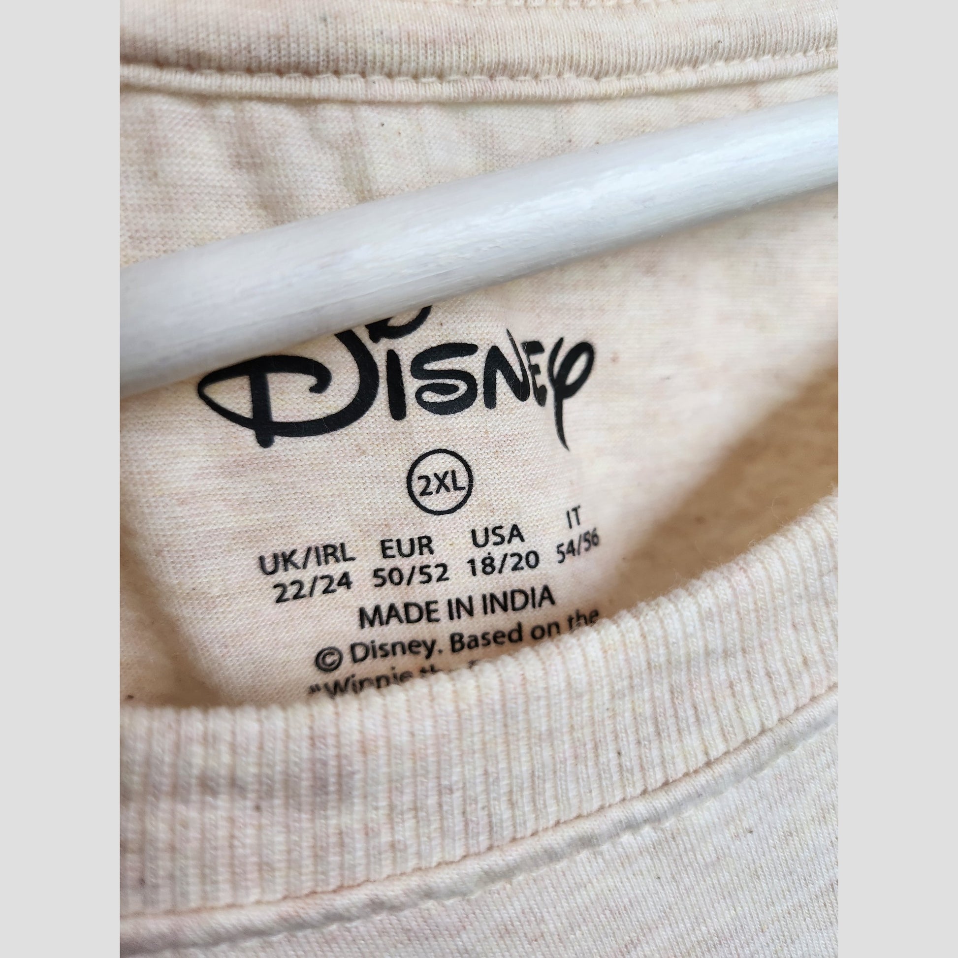 Close-up of Primark's Upcycled Breastfeeding & Pumping Cream Winnie the Pooh t-shirt label, size 2XL (UK22/24), made in India, with multi-region sizing in black—part of our preloved maternity wear collection.