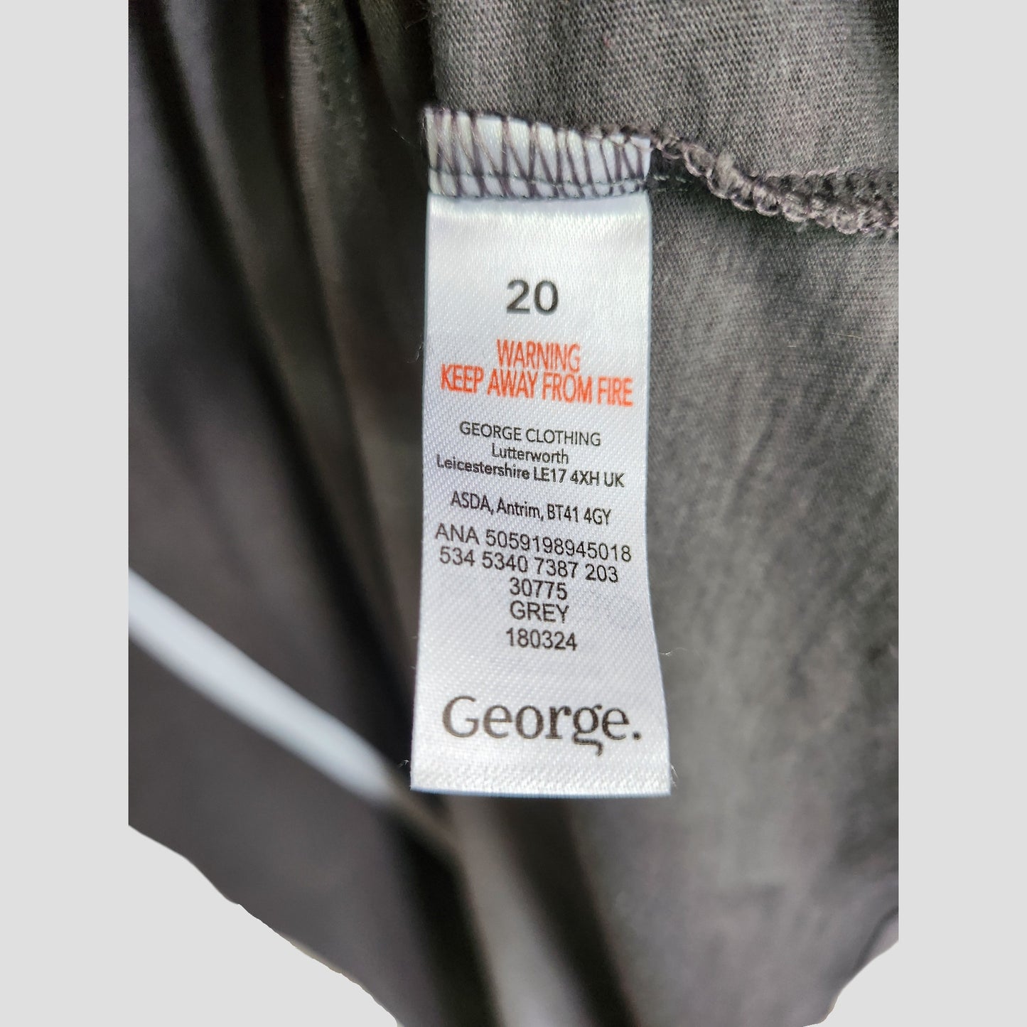 A close-up of a white label on grey fabric from George, reading “20, WARNING KEEP AWAY FROM FIRE,” features size UK20 info on the Upcycled Sunshine Moomin Nursing & Pumping t-shirt—ideal for sustainable maternity fashion lovers.
