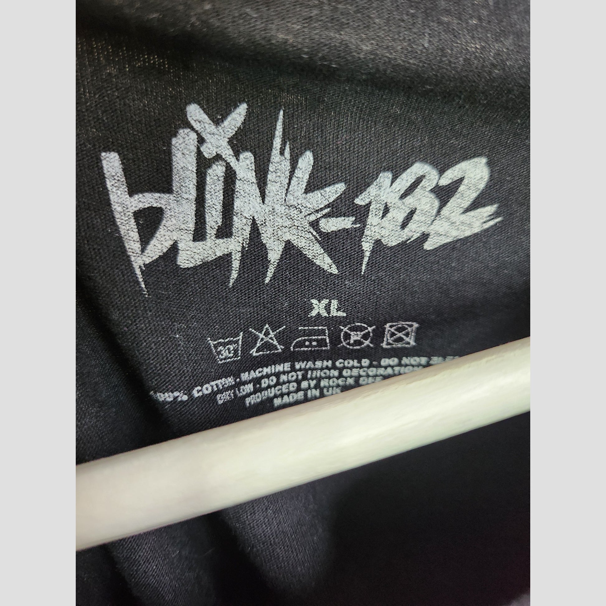 Black/Pink Blink 182 upcycled nursing & pumping t-shirt (XL) label features the band’s logo in white, size/isntructions/material details—an eco-friendly maternity option—partially hidden by a white hanger. Brand: Blink 182.