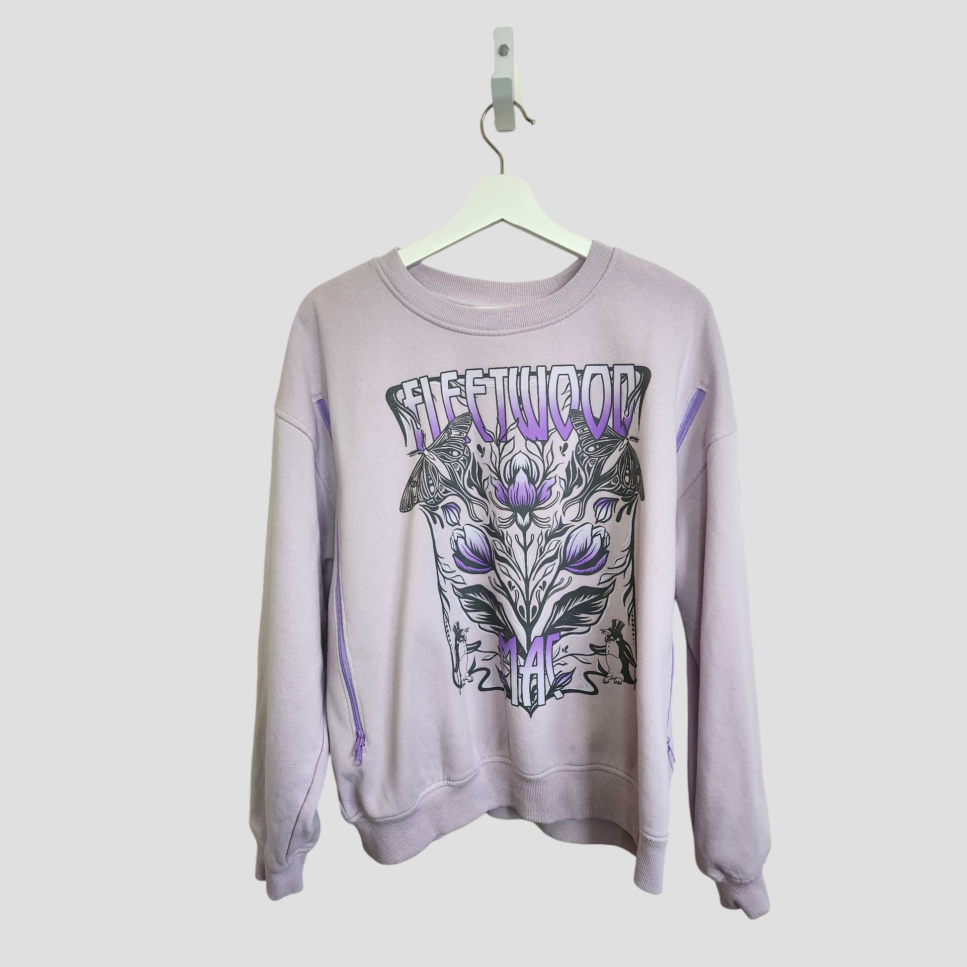 A lilac H&M upcycled breastfeeding & pumping Fleetwood Mac band sweatshirt (L, 100135) hangs on a white hanger, showcasing bold black and purple graphics with floral and geometric designs—eco-friendly, stylish, and affordable.