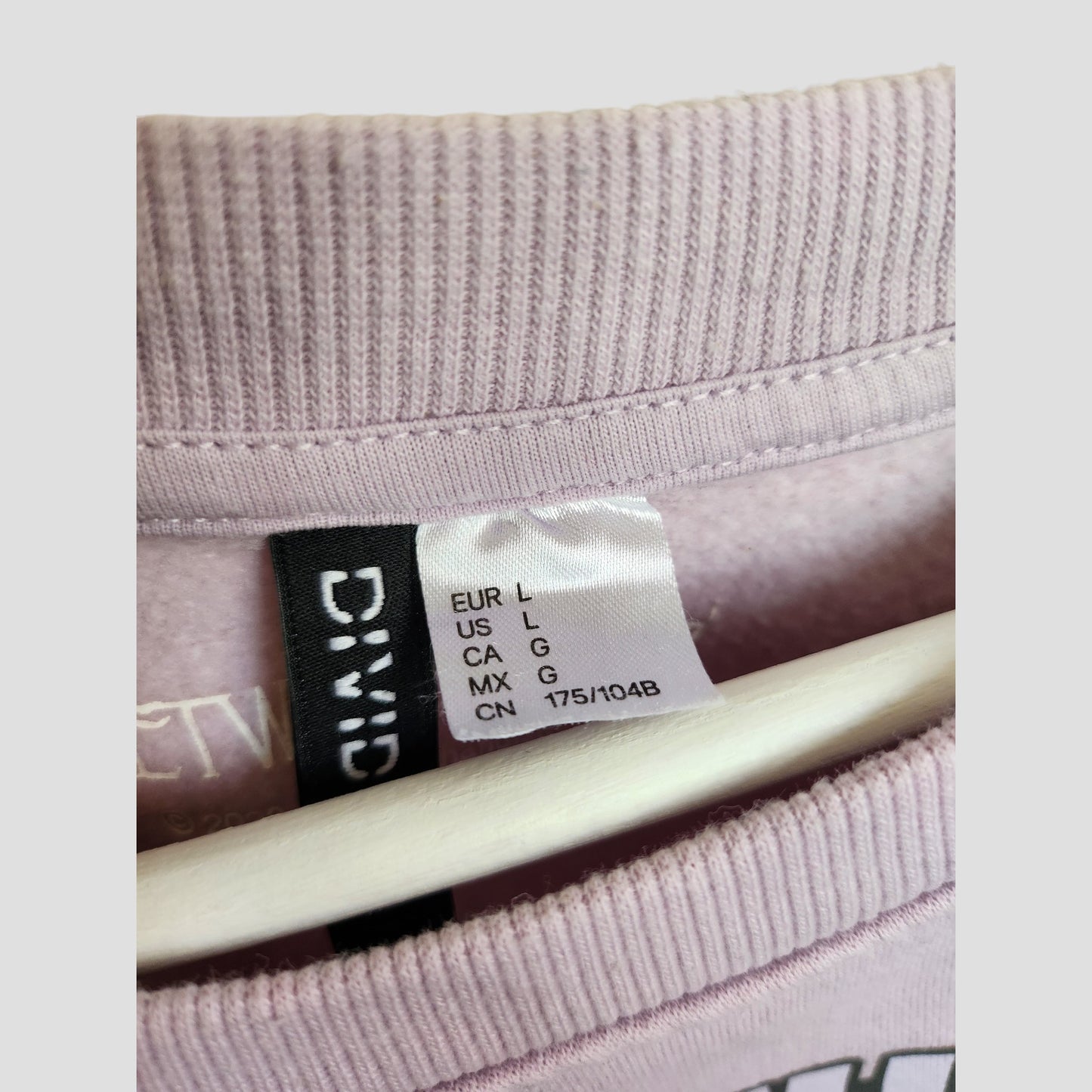 Close-up of an H&M label on a lilac Upcycled Breastfeeding & Pumping Fleetwood Mac Band Sweatshirt (L), showing sizes: EUR L, US L, CA G, MX G, CN 175/104B. The eco-affordable garment hangs on a white hanger with "DIVIDED" visible.