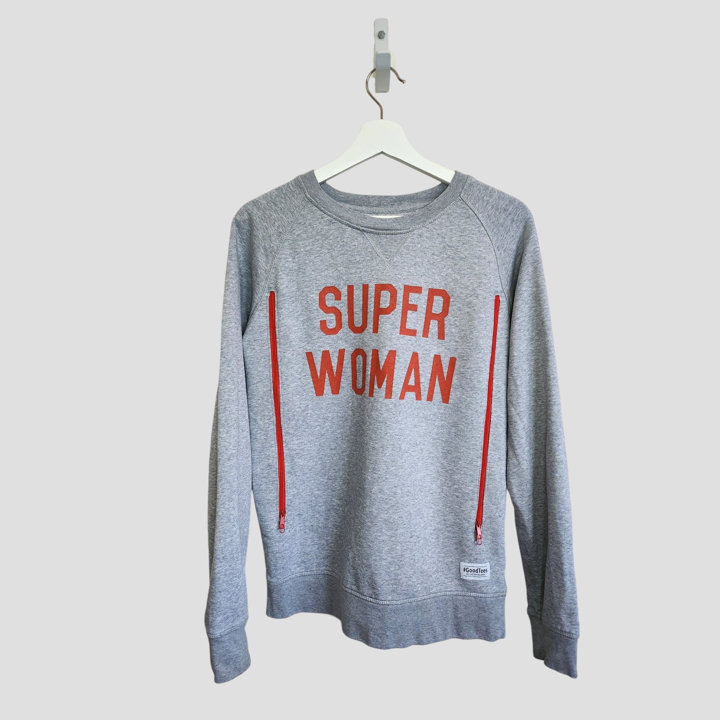 The Selfish Mother Upcycled Breastfeeding & Pumping Grey/Red SUPER WOMAN Sweatshirt (M, 100155) hangs on a white hanger, featuring bold red lettering and two vertical red zippers against a plain background.