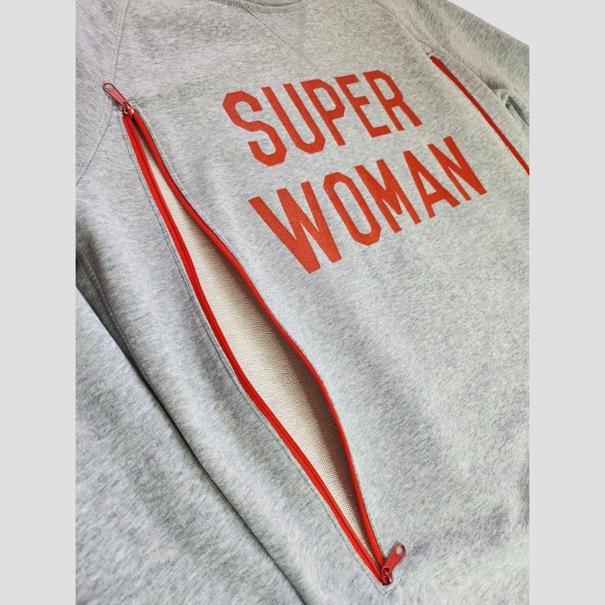 The Selfish Mother Upcycled Breastfeeding & Pumping Grey/Red SUPER WOMAN Sweatshirt (M) features bold red lettering, diagonal red zippers for easy nursing or pumping, and eco-friendly style for practical moms.