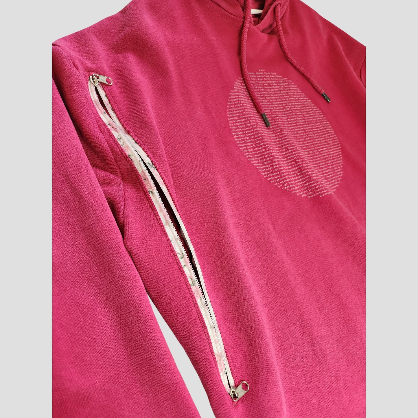 The Human Milk Upcycled Breastfeeding Hoodie L features a bright pink design, a large textured circle on the chest, and a diagonal white zipper on the sleeve that opens to reveal inner fabric—eco-friendly maternity fashion for nursing or pumping.