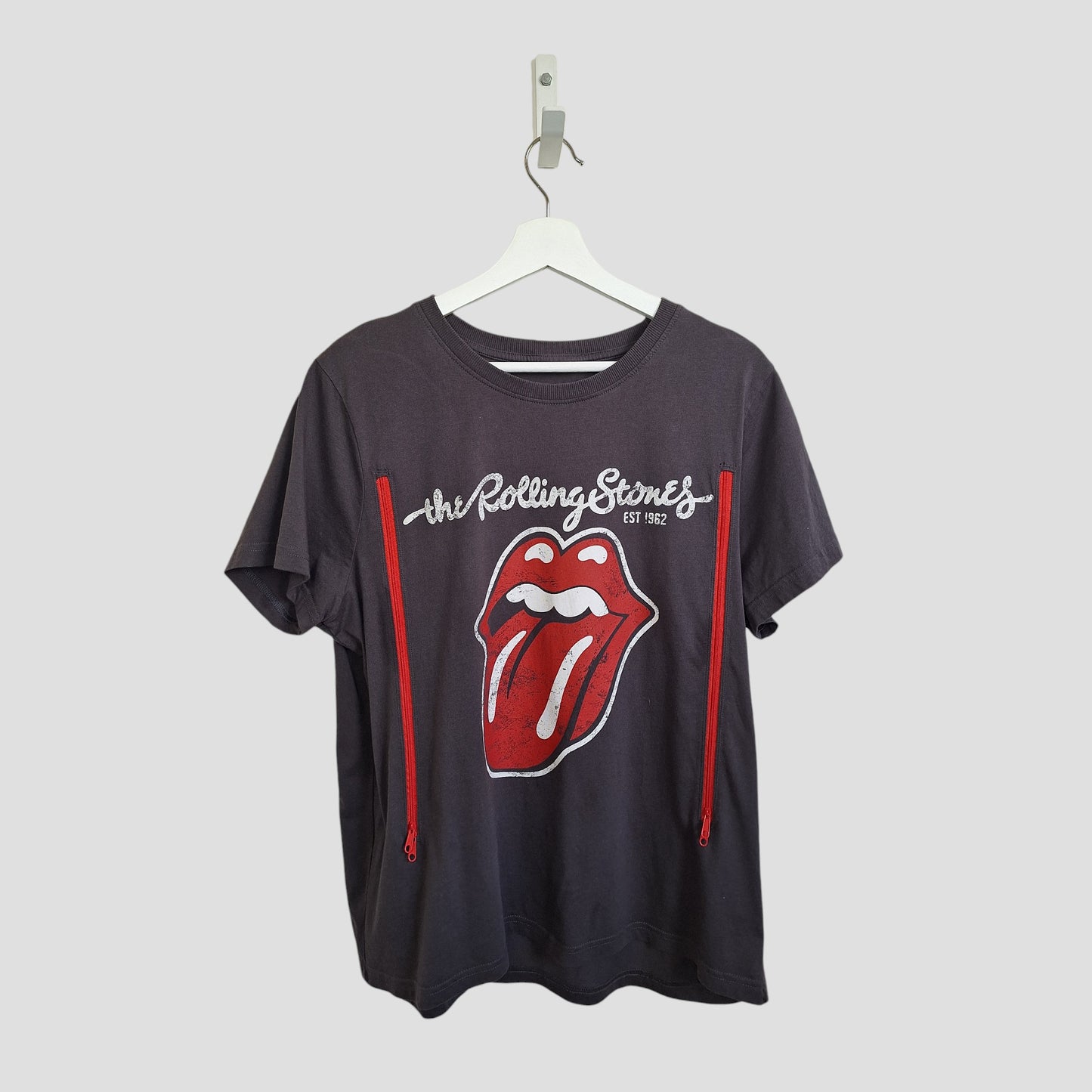 A grey upcycled Primark Rolling Stones nursing & pumping band t-shirt (L), featuring the band's name and logo, made from preloved clothing, hangs on a white hanger against a plain light background.