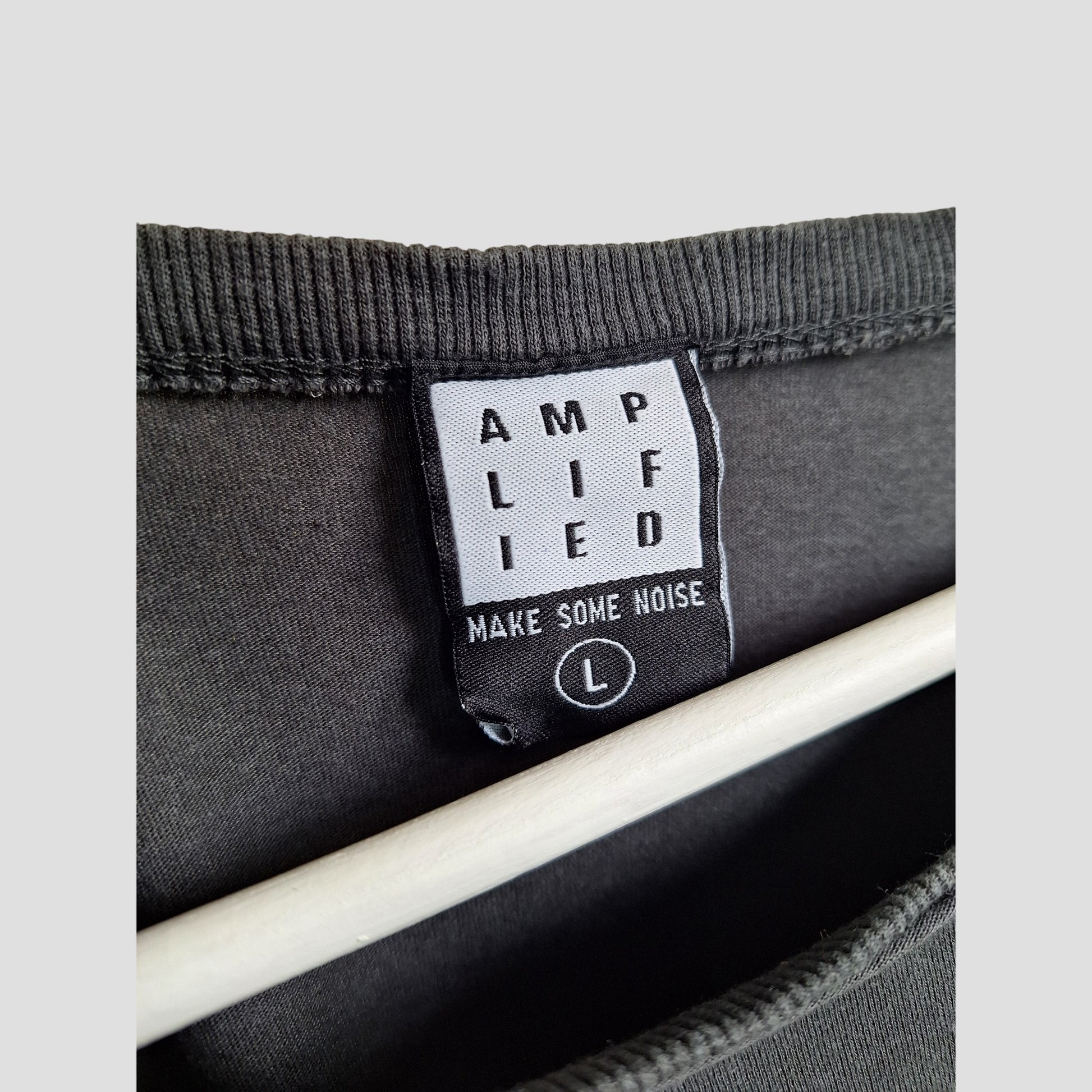 Close-up of a black AMPLIFIED label reading "MAKE SOME NOISE" with an L for size inside an upcycled breastfeeding & pumping grey/yellow Iron Maiden band t-shirt (L, 100352), shown on black fabric and a wooden hanger.