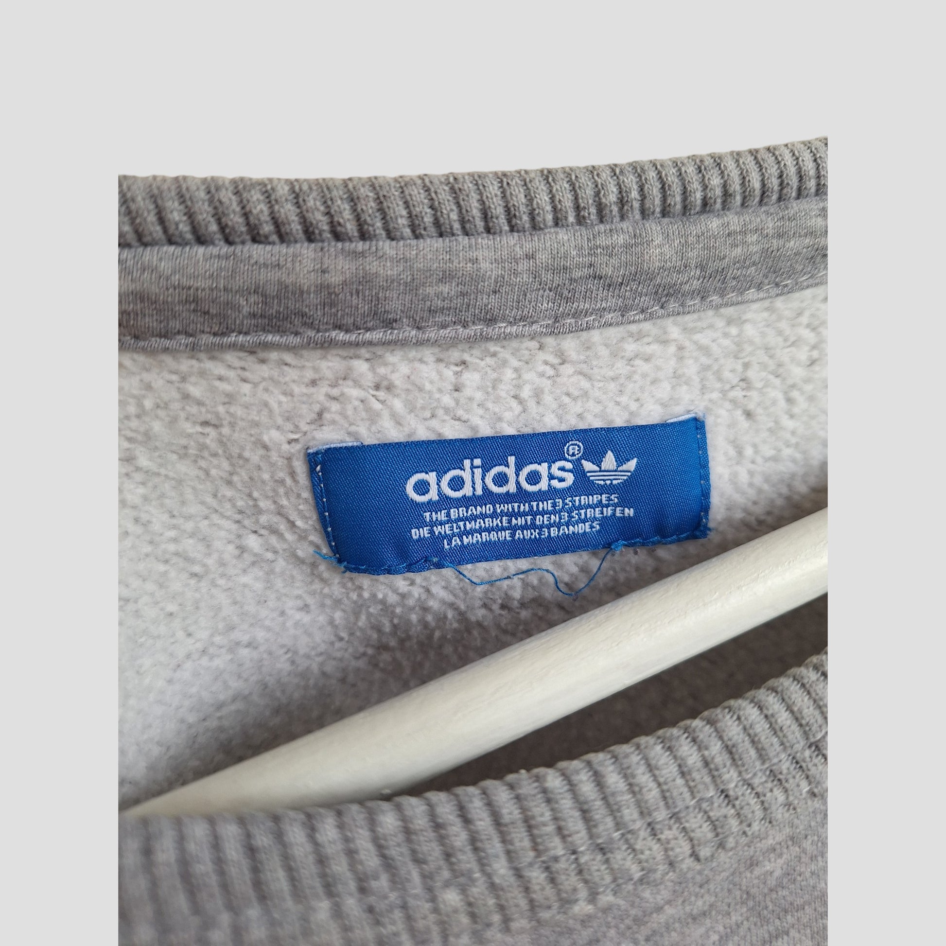 Close-up of a blue Adidas tag sewn into an upcycled grey fleece-lined Adidas Breastfeeding & Pumping Sweatshirt (L, UK14), logo and text visible on the preloved garment, which is hanging on a white hanger.
