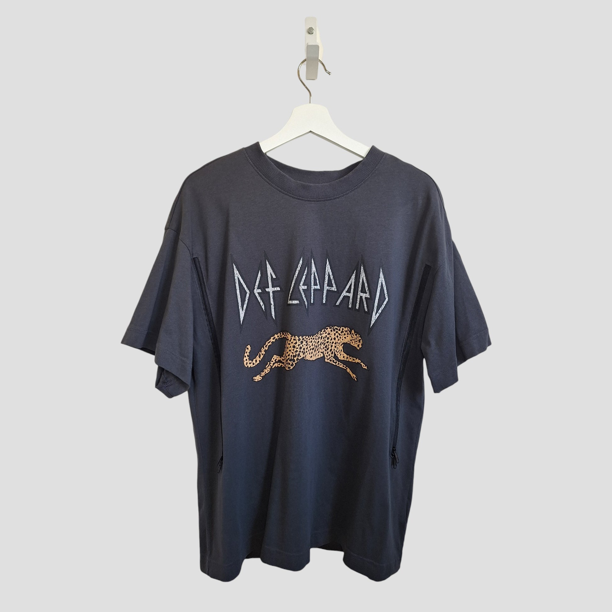 An upcycled grey Def Leppard band T-shirt by H&M hangs on a white hanger, combining eco-friendly nursing and pumping clothing with edgy band graphics for a unique, conscious look.